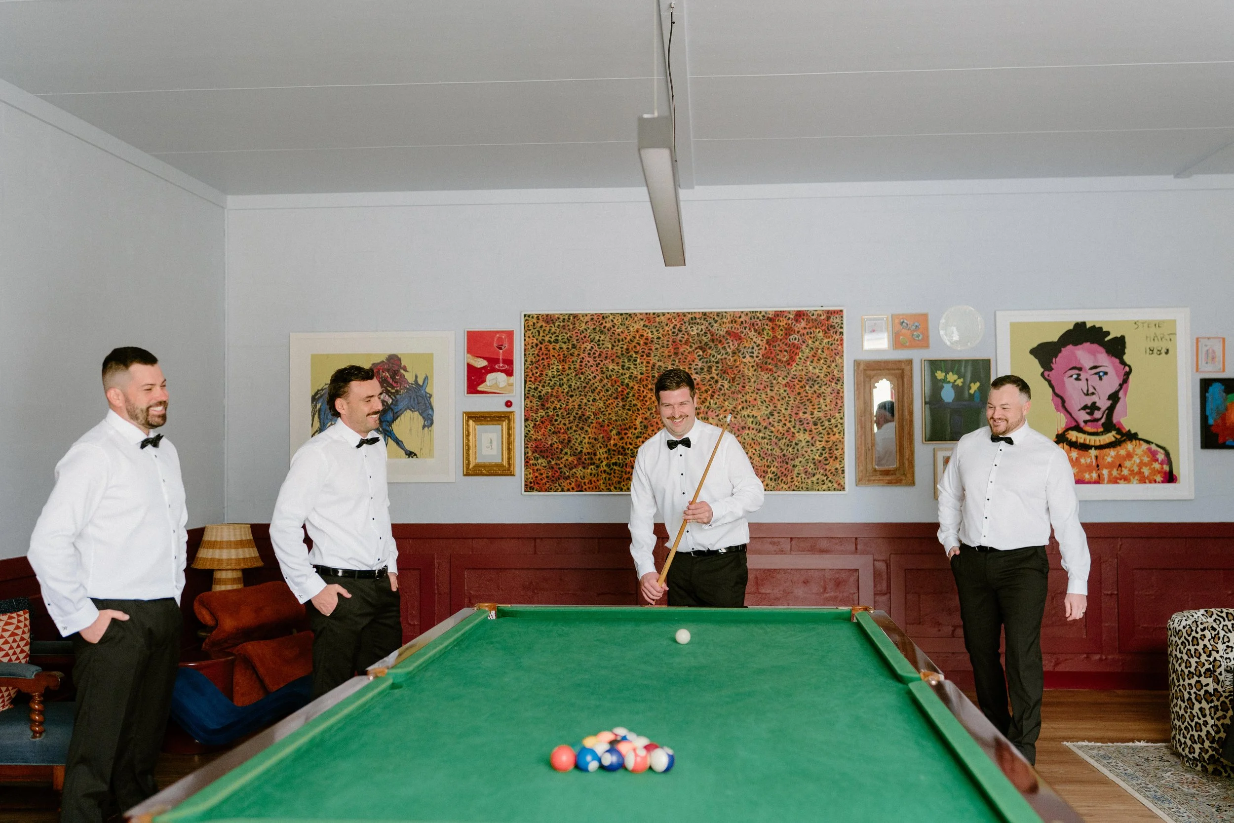 Five men in white shirts and black bow ties playing pool in a room decorated with colorful artwork.