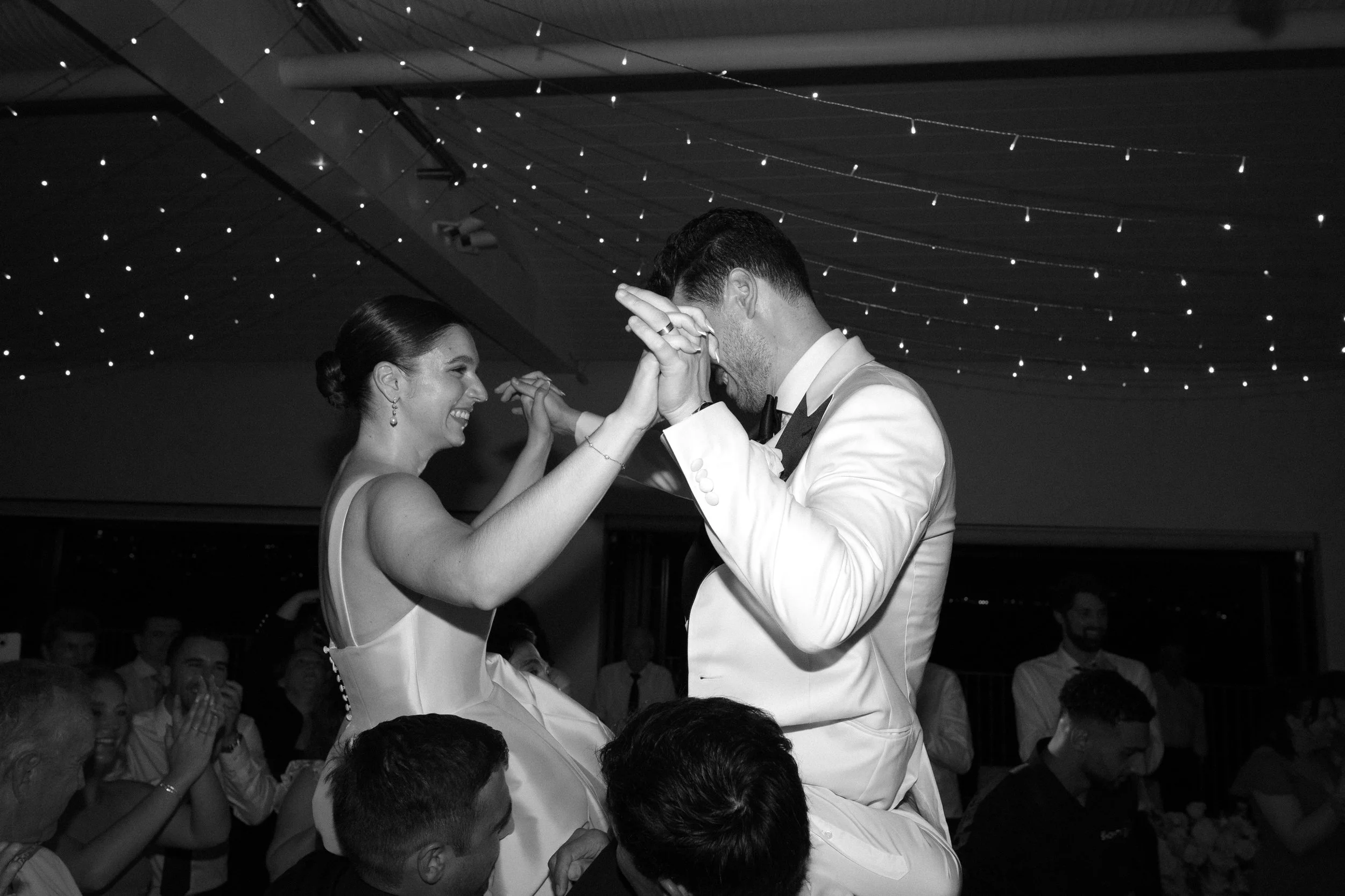 A bride and groom are dancing at their wedding reception surrounded by smiling guests, with string lights overhead.