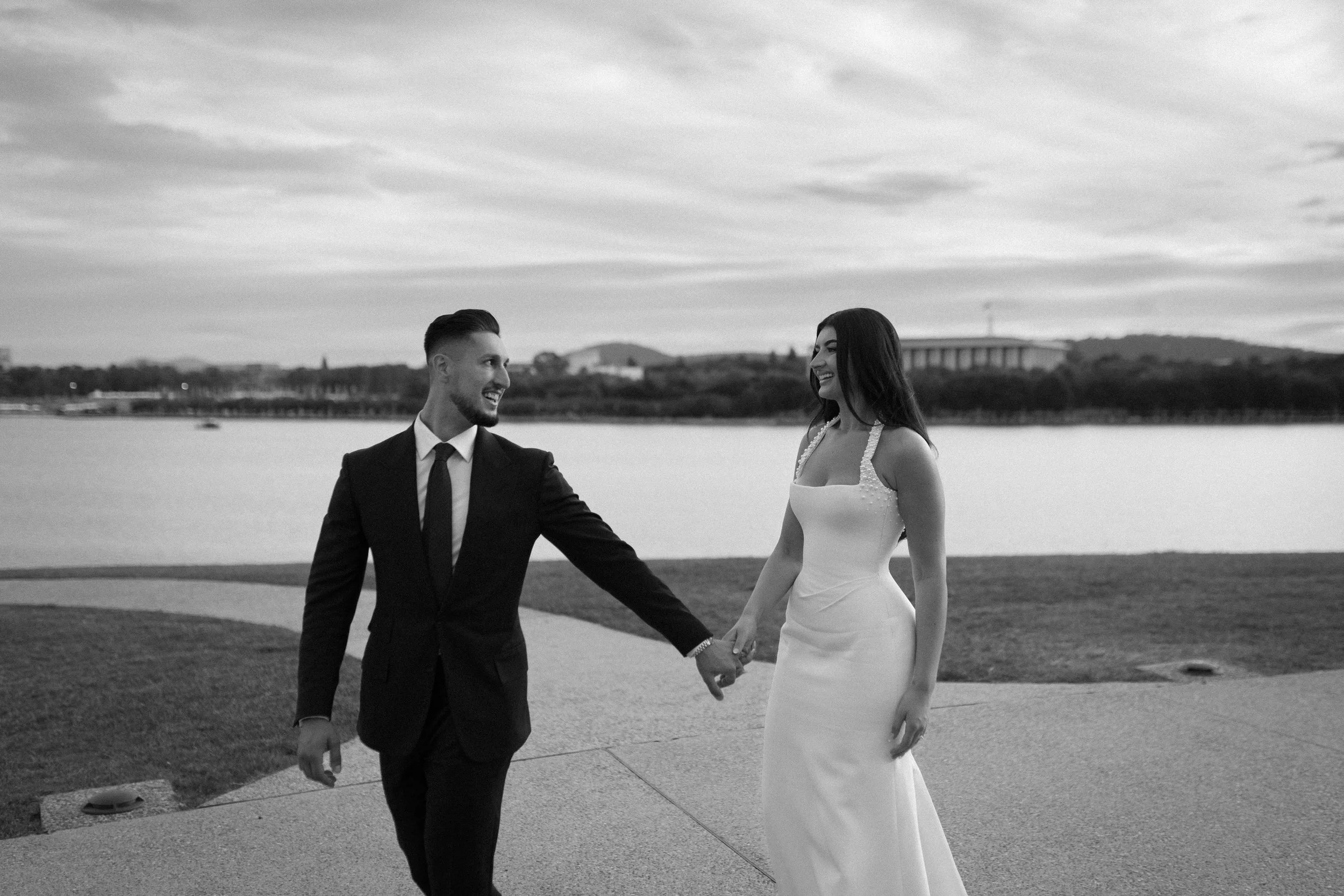 A black and white photo of a bride and groom holding hands and smiling at each other outdoors near a body of water with a bridge and hills in the background.