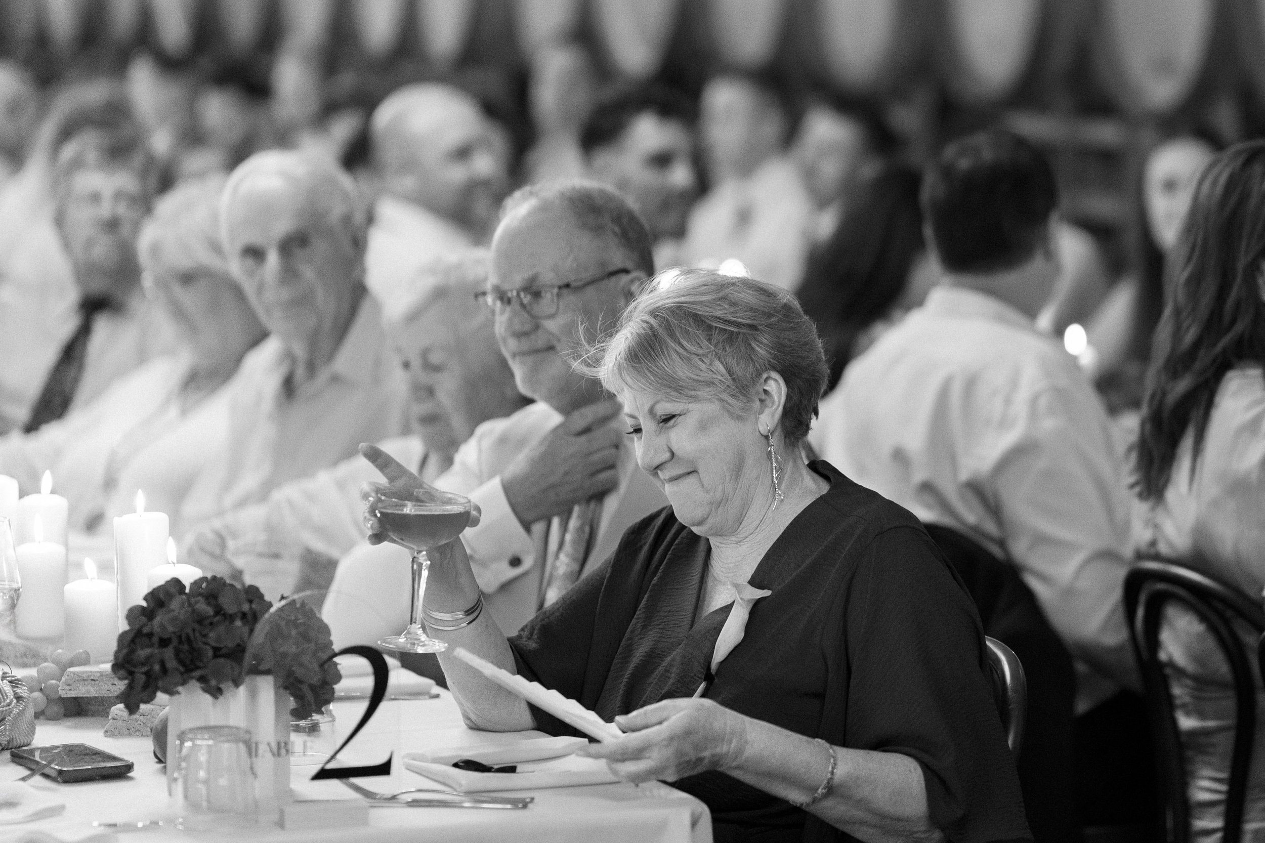 An elderly woman sitting at a table during a formal event, holding a cocktail glass in one hand and reading a piece of paper in the other, surrounded by candles and floral centerpieces.