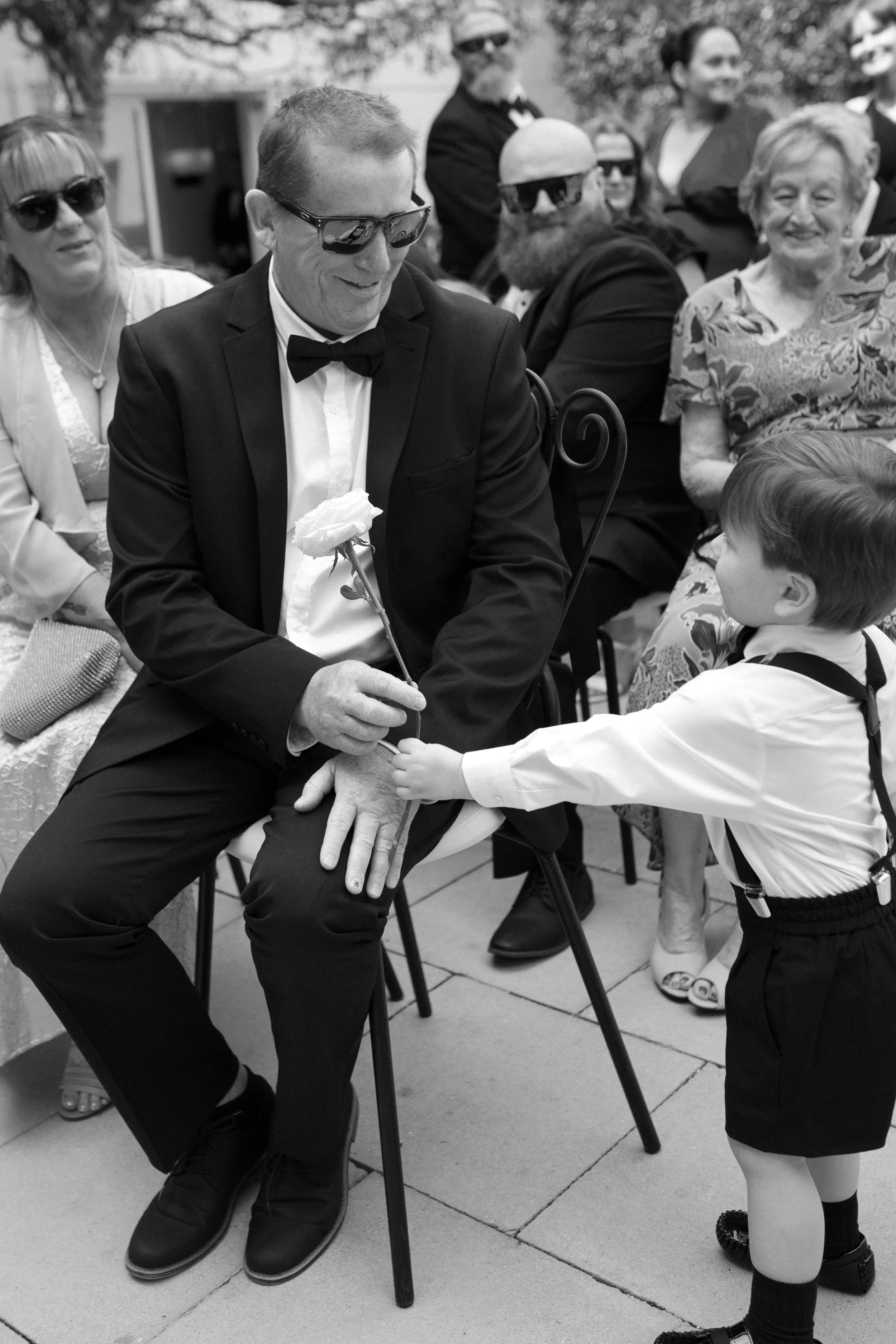 A man in a tuxedo and sunglasses is seated, holding a rose, as a young boy in a white shirt and black shorts offers him the flower during a social event.