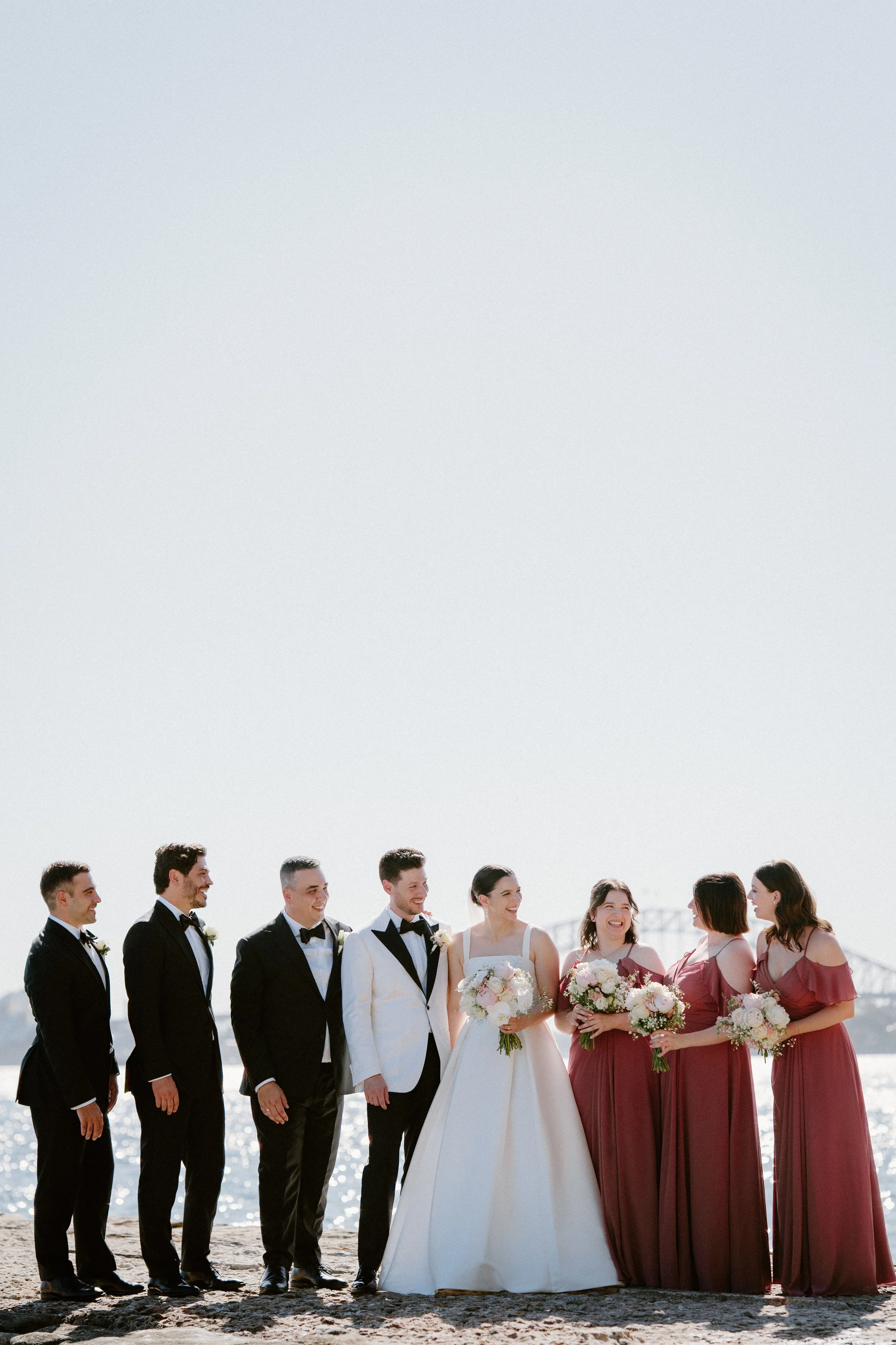 A wedding party consisting of six men in tuxedos and five women in bridesmaid dresses, standing on a beach with water and a bridge in the background, smiling and holding bouquets.