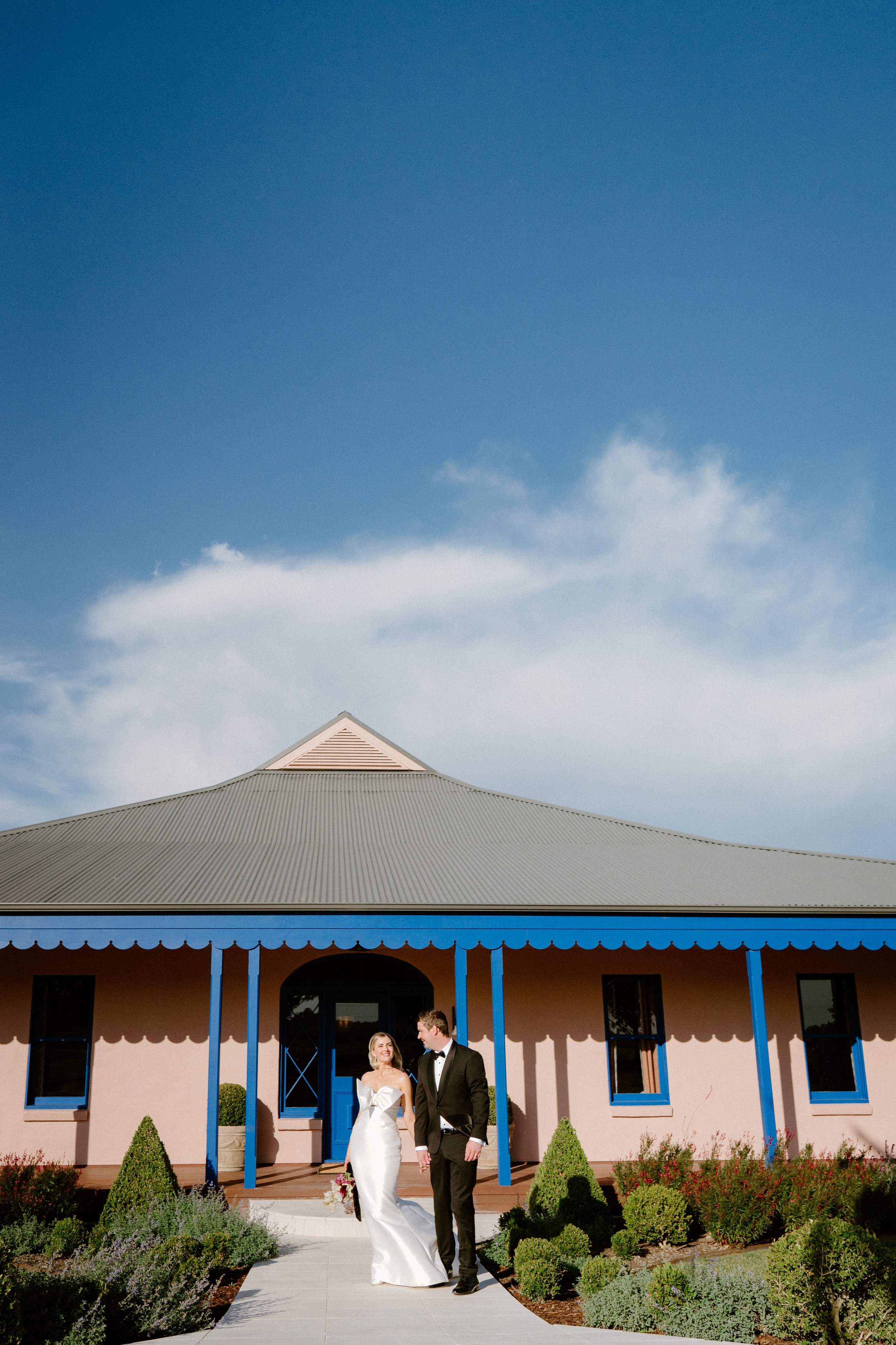 The Sienna, Hunter Valley Wedding Venue, Wedding Photos. A bride and groom holding hands while walking outside a pink building with blue trim on a sunny day.