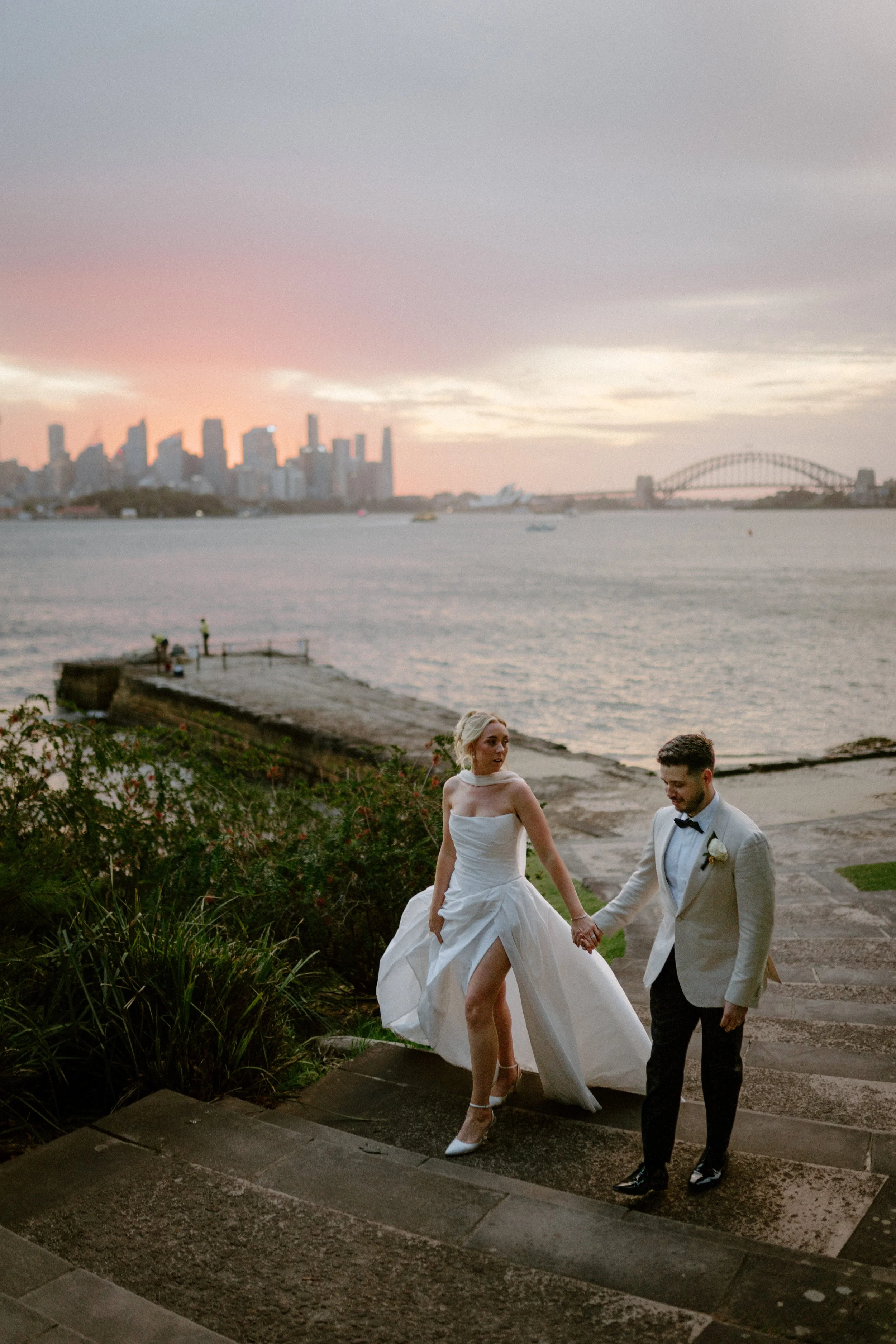 A bride and groom walking hand in hand on a staircase near the water at sunset, with the Sydney Opera House and Harbour Bridge visible in the background.