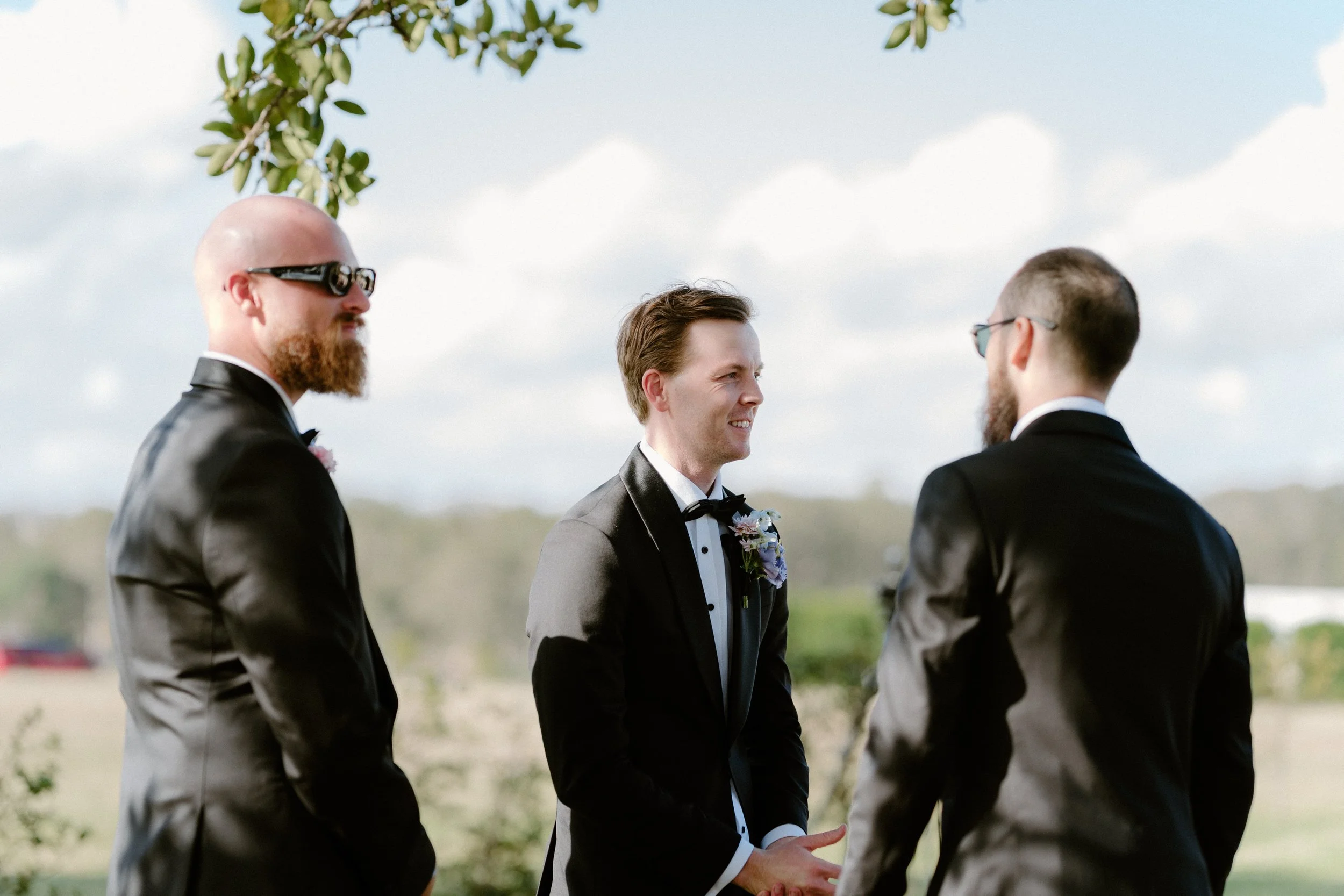 Three men dressed in tuxedos and wearing sunglasses, standing outdoors with a cloudy sky and some greenery in the background.