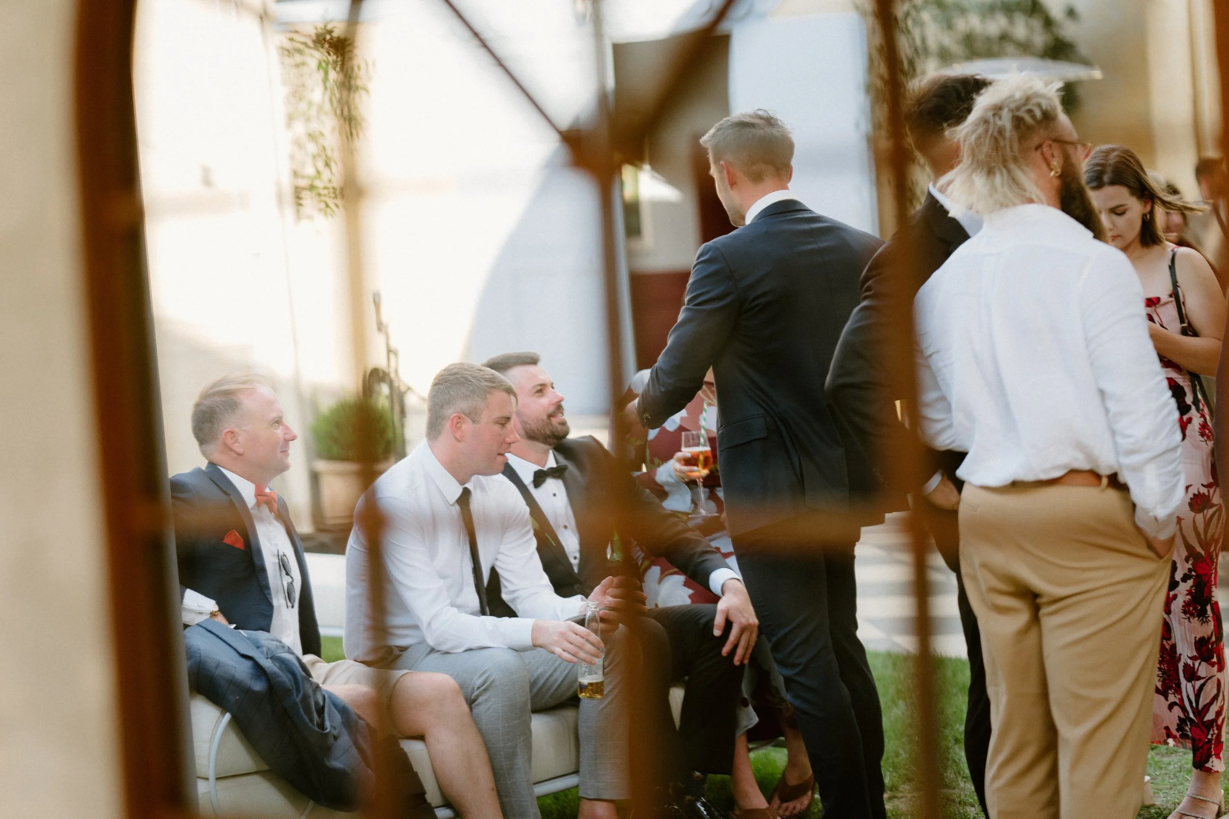 Group of well-dressed people socializing outdoors, with some sitting on a couch and others standing, during a formal or semi-formal event.