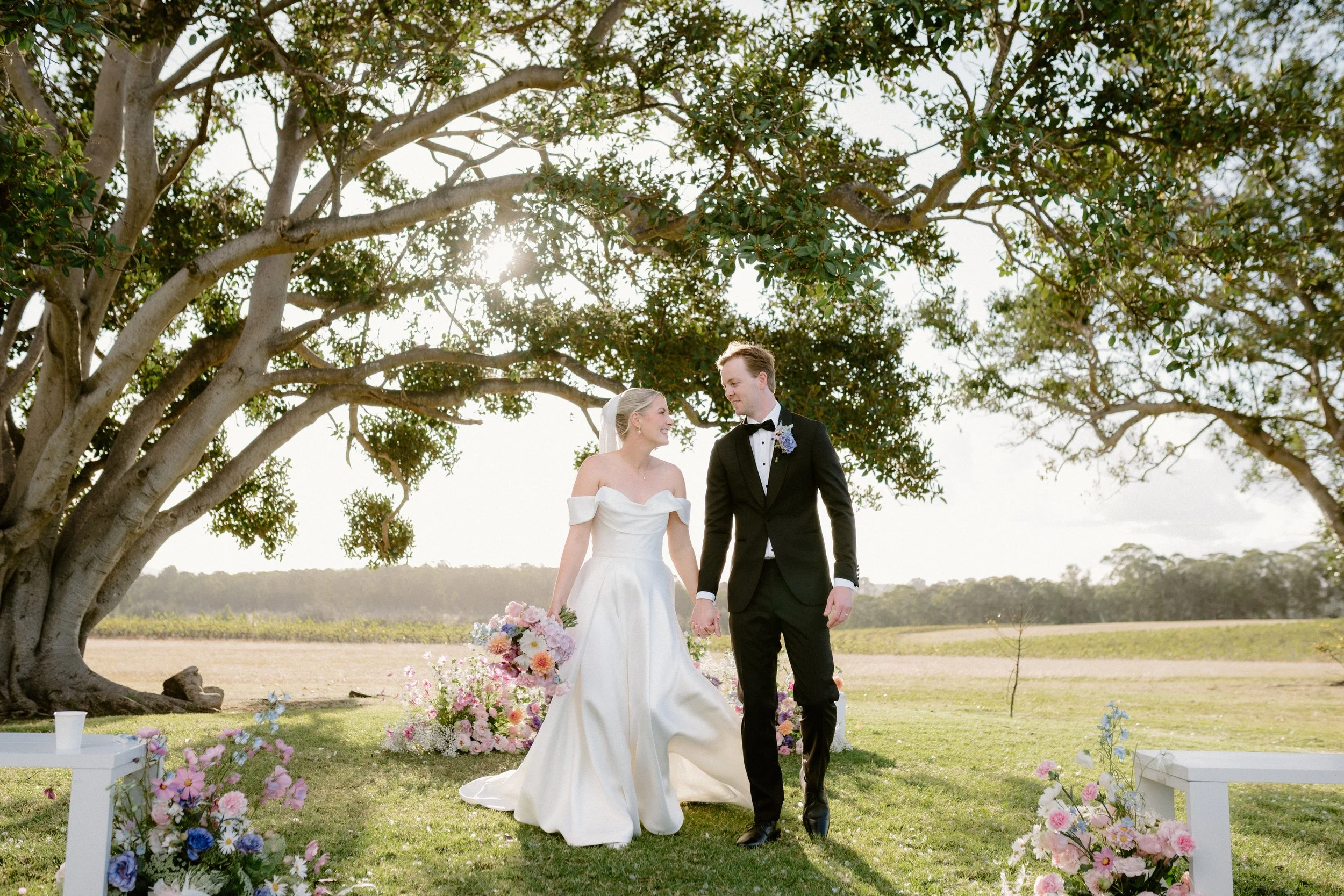 A bride and groom walking hand in hand outdoors during their wedding ceremony, surrounded by flowers under a large tree with sunlight shining through the leaves. Bimbadgen Palmers Lane Wedding, Hunter Valley