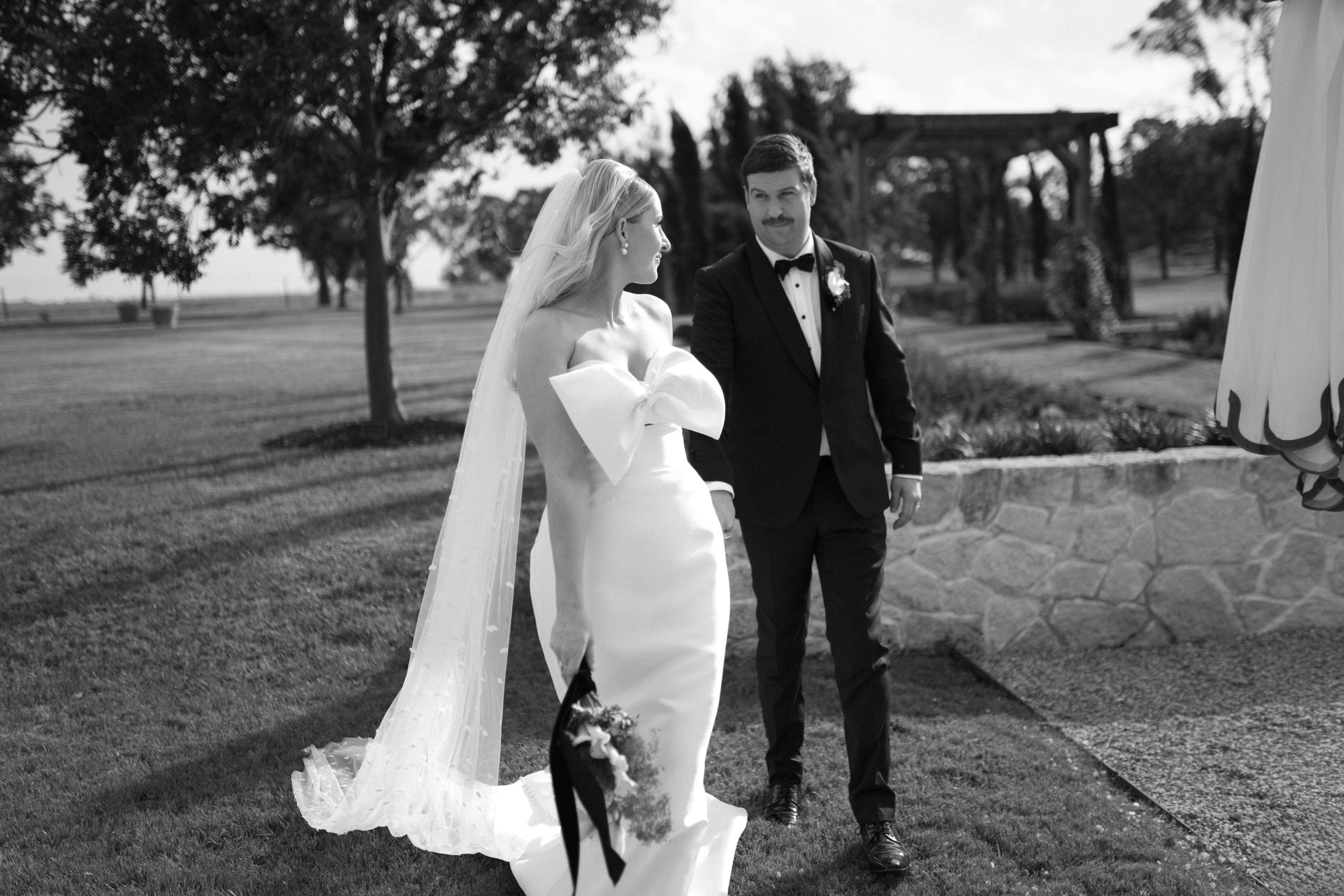 Black and white photo of a bride and groom walking hand in hand outdoors, trees and a stone wall in the background.