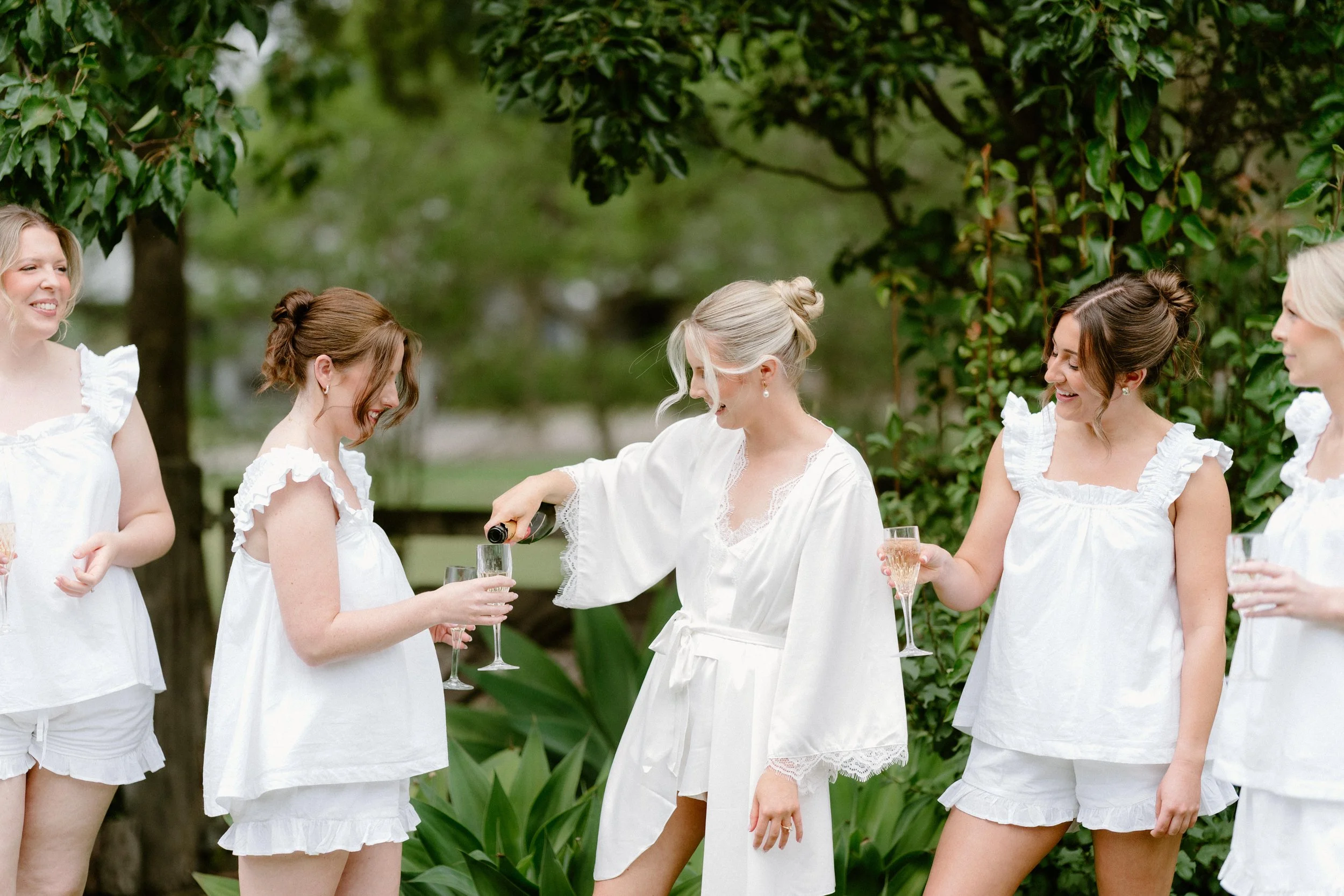 A group of women dressed in white, celebrating outdoors with champagne glasses, with one pouring champagne, surrounded by greenery.