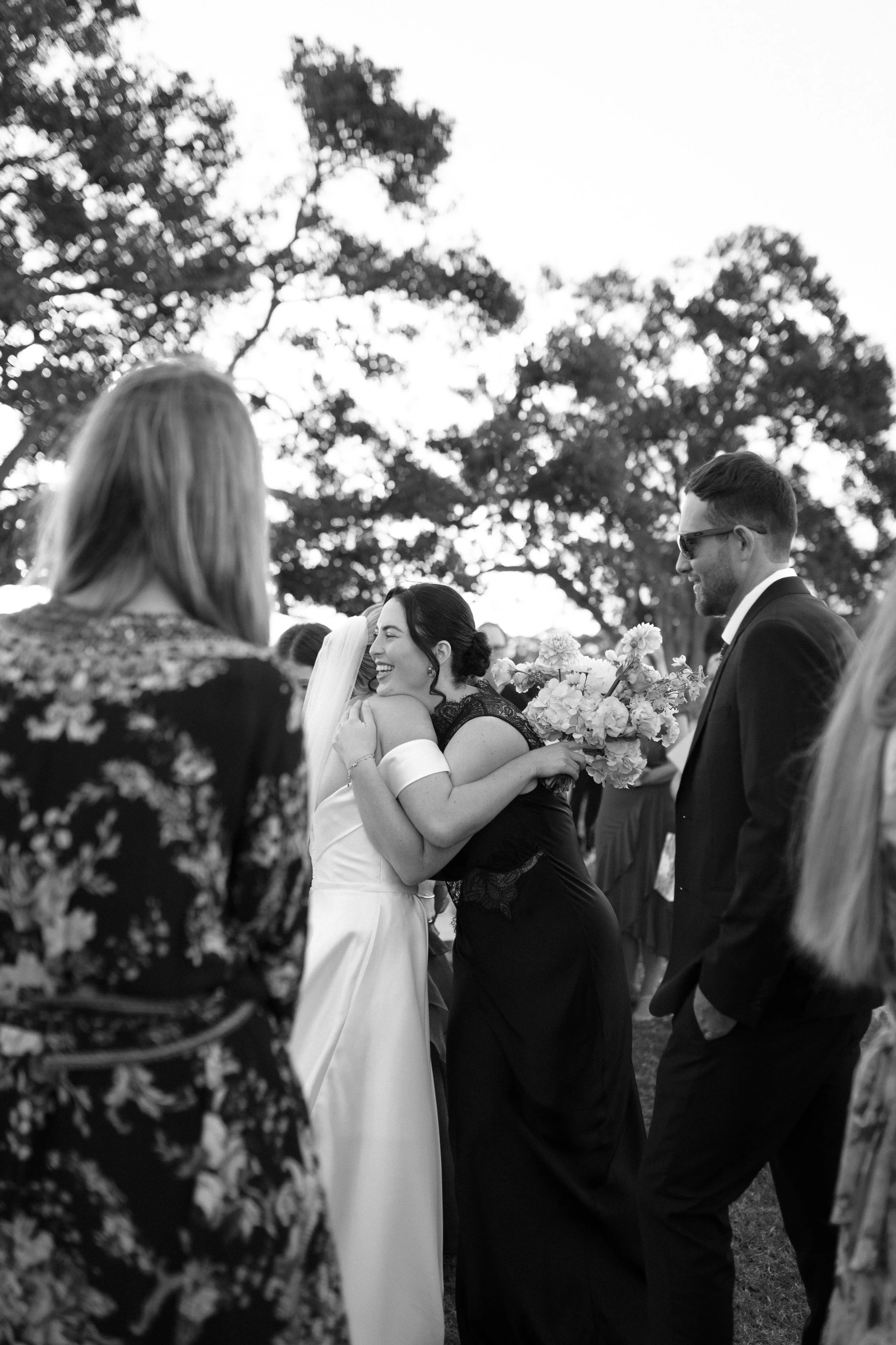 Black and white photo of a wedding ceremony outdoors, with a bride hugging a guest, surrounded by well-dressed attendees, tall trees in the background, and the bride holding a bouquet of flowers.