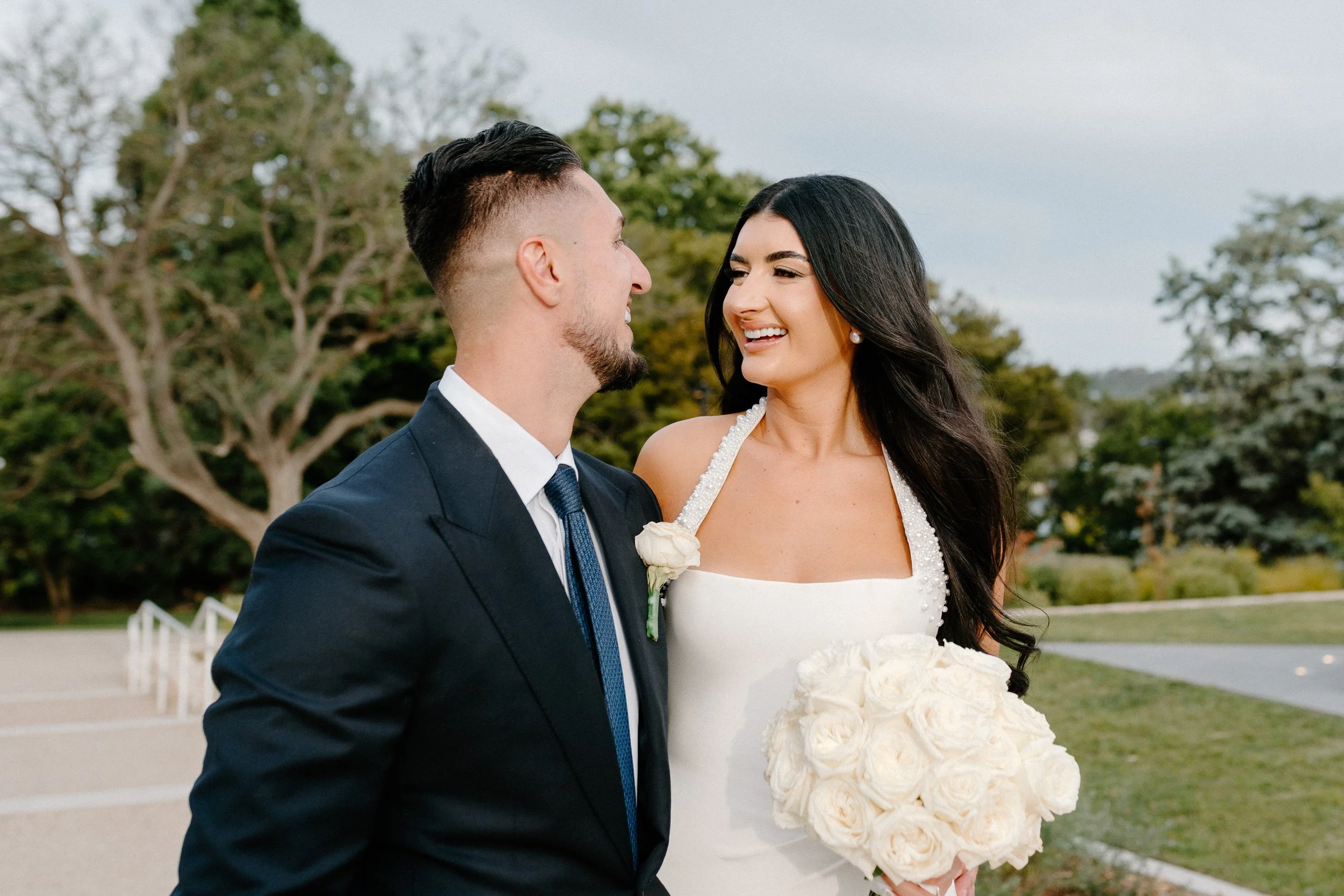 A bride and groom standing outdoors, smiling at each other, with trees and a park in the background.