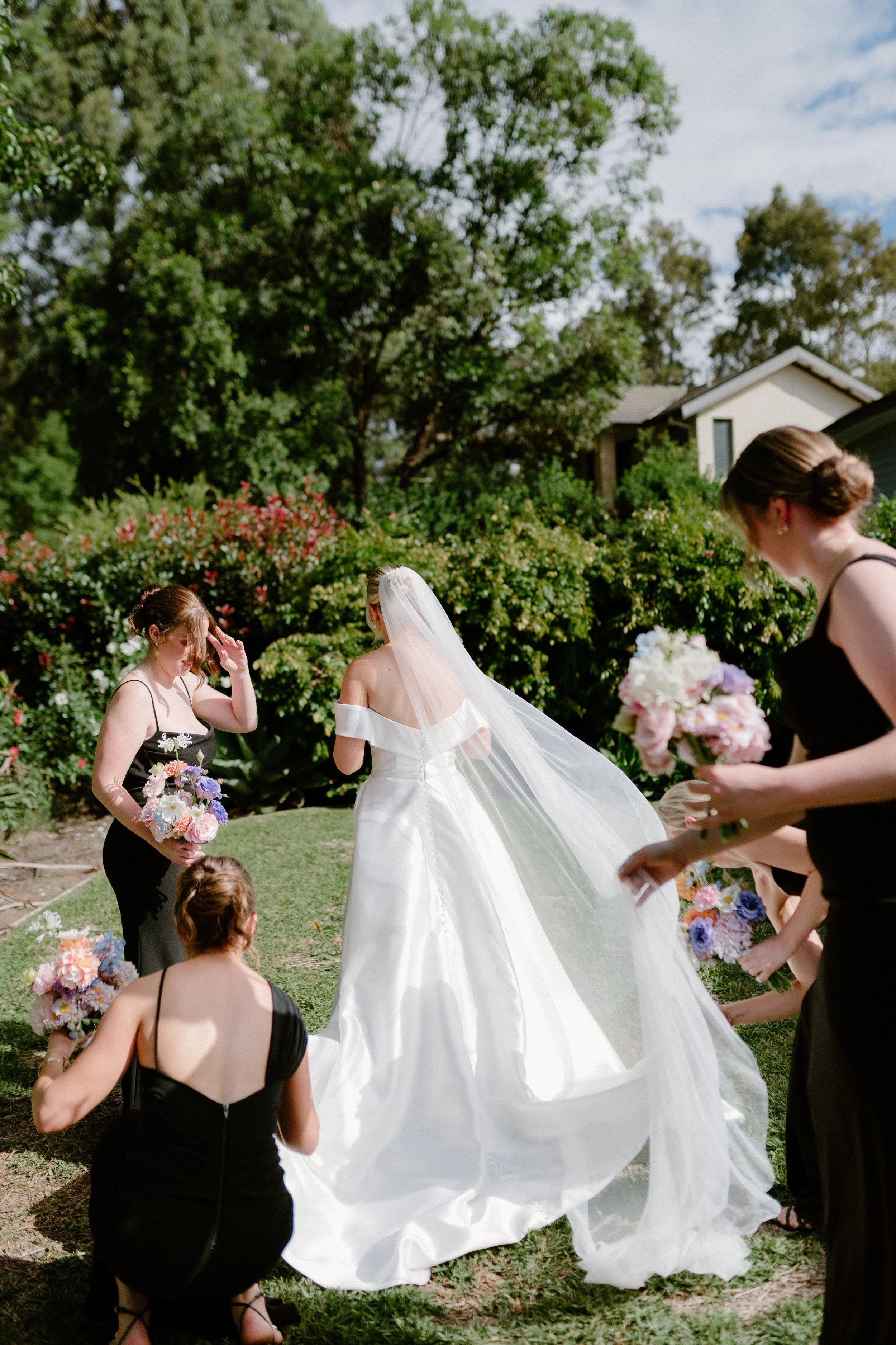 A bride in a white wedding dress and veil standing outdoors surrounded by bridesmaids in black dresses holding colorful bouquets of flowers while they prepare for a wedding ceremony. Bimbadgen Palmers Lane Wedding, Hunter Valley