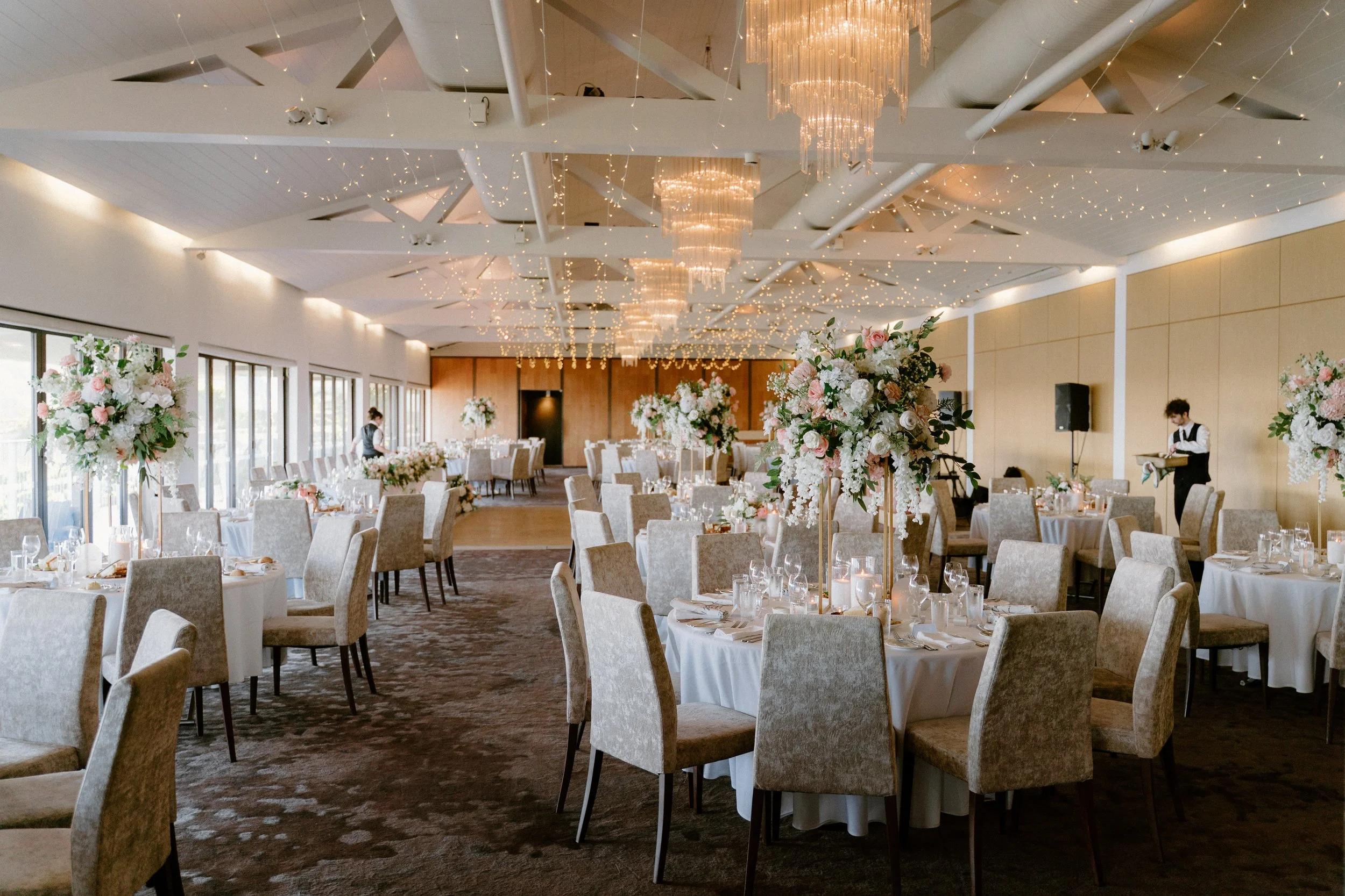 Elegant wedding reception hall with round tables adorned with white tablecloths, tall floral centerpieces of pink and white flowers, surrounded by beige upholstered chairs. The ceiling features string lights and chandeliers, with large windows along 