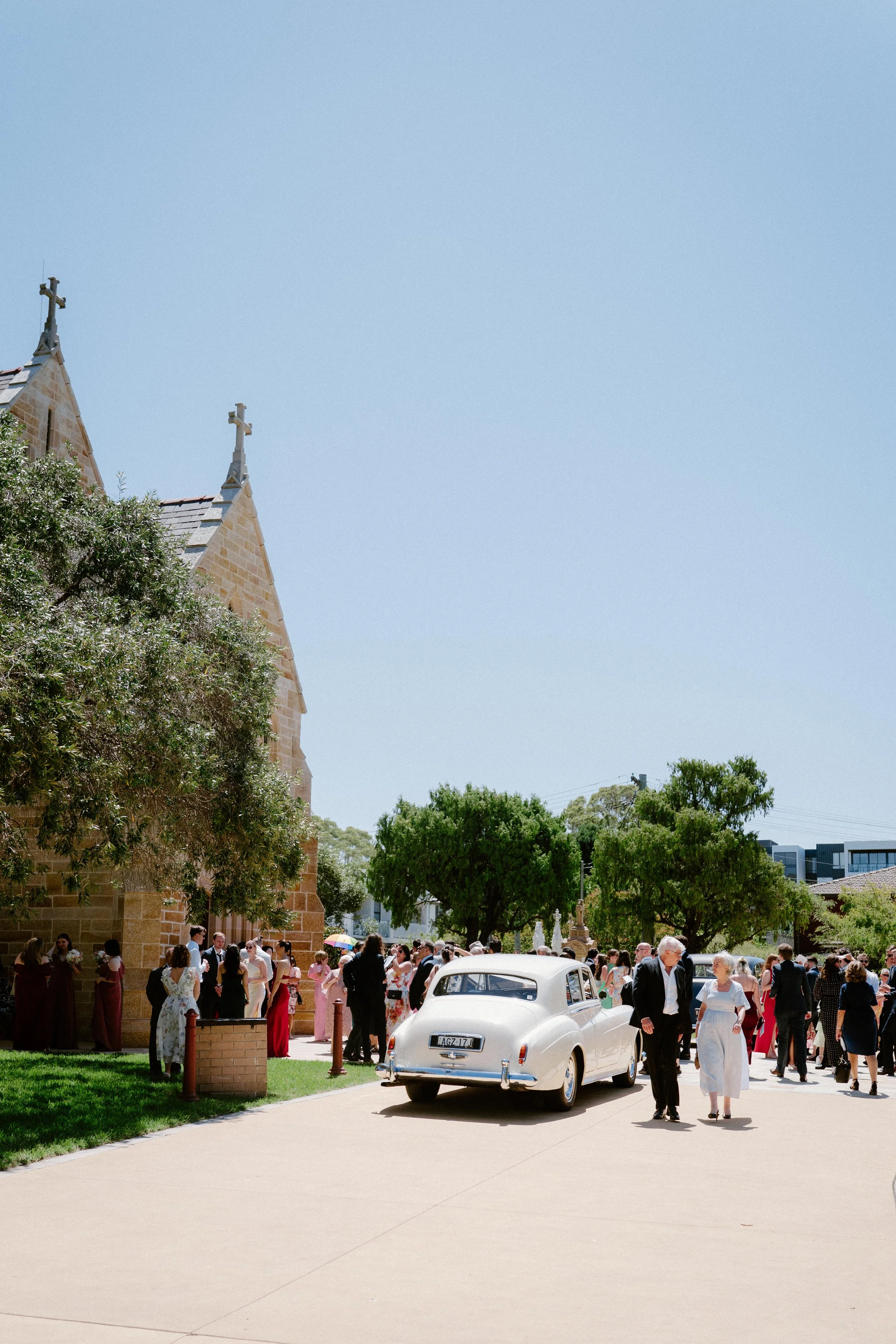 A wedding celebration outside a stone church with guests dressed in formal attire, vintage white car parked nearby, and green trees under a clear blue sky.