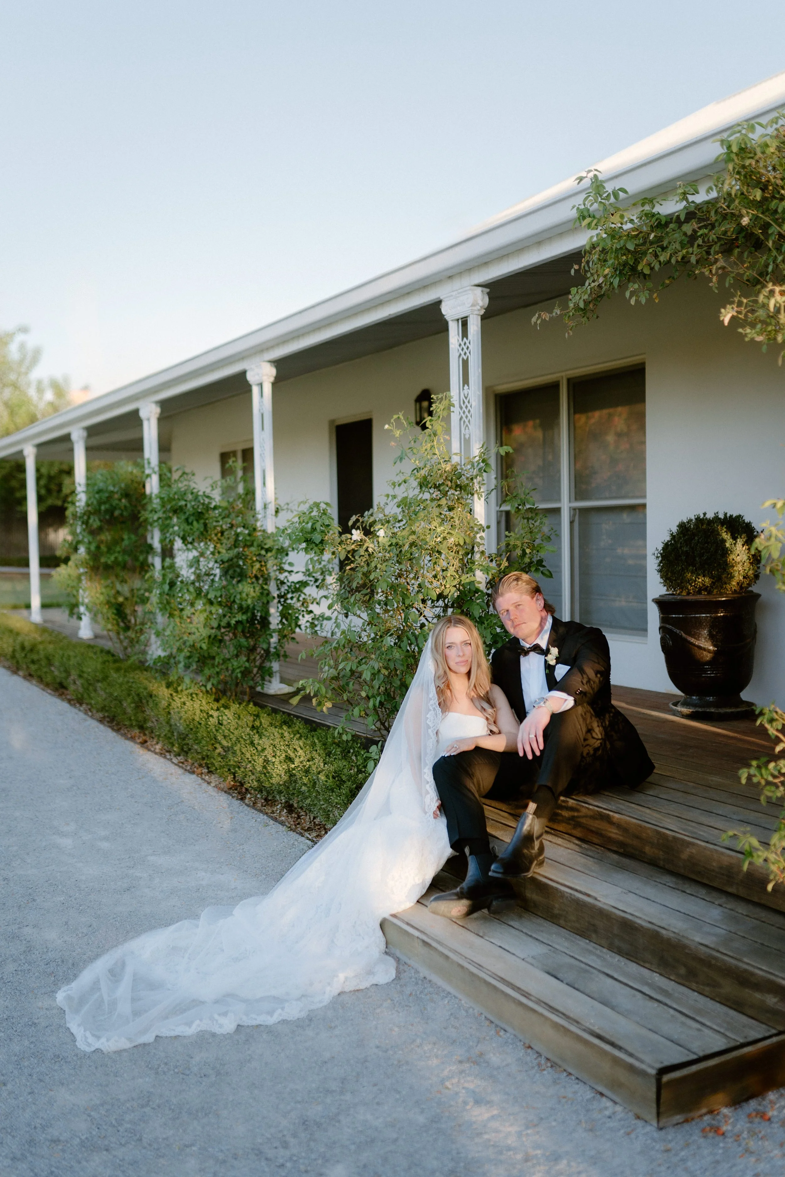 A bride in a white wedding dress with a long train and veil, and a groom in a black tuxedo, sitting together on wooden steps outside of a house with white exterior walls, surrounded by green bushes and potted plants.