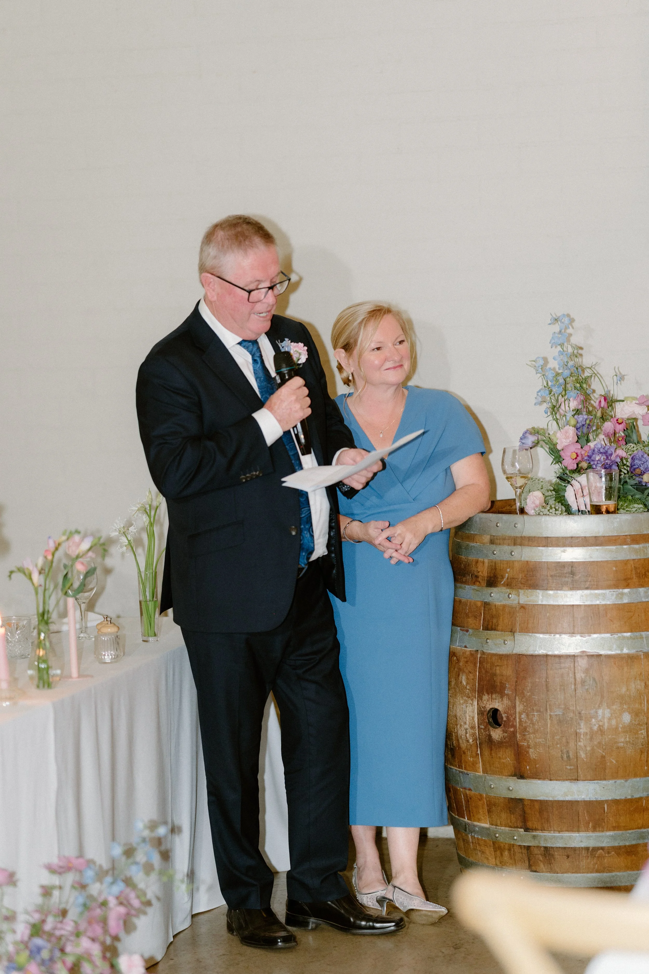 A man in a black suit, white shirt, and blue tie, with glasses, is speaking into a microphone. A woman in a blue dress stands beside him, listening with a smile. The setting appears to be a celebratory event with floral decorations and a large wooden