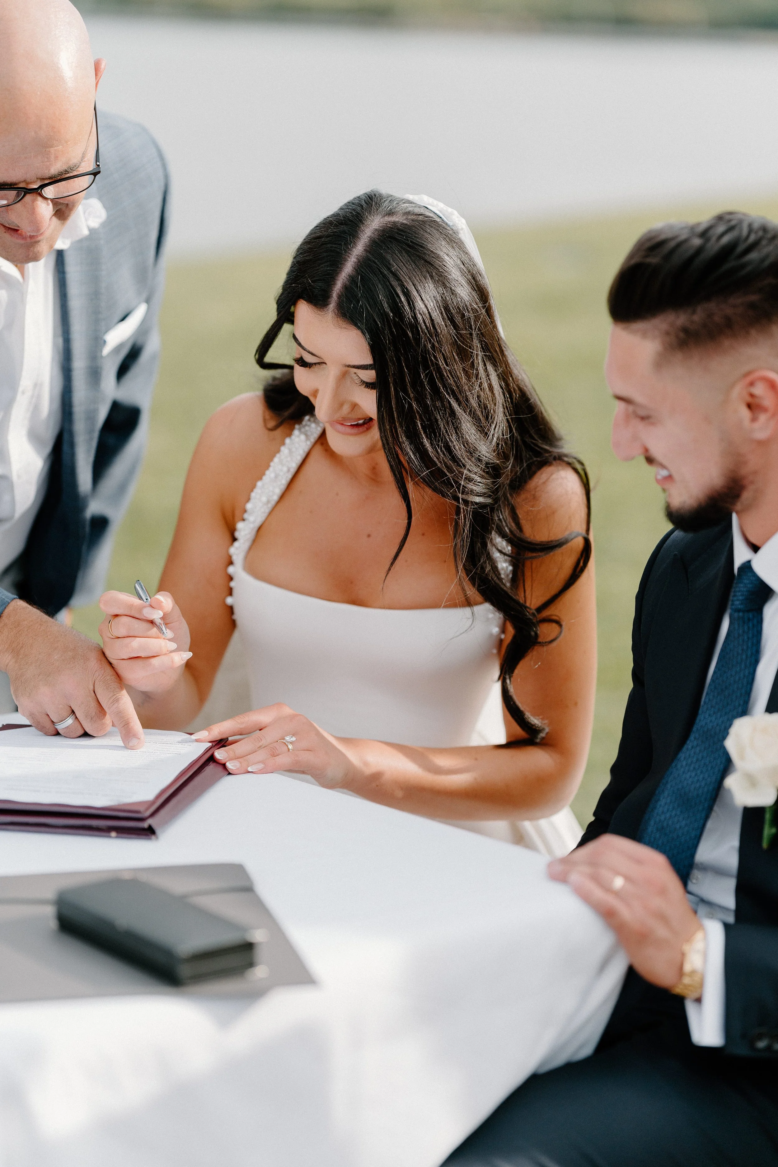 A bride signing a marriage certificate at her wedding ceremony outdoors, with a groom and a person officiating.