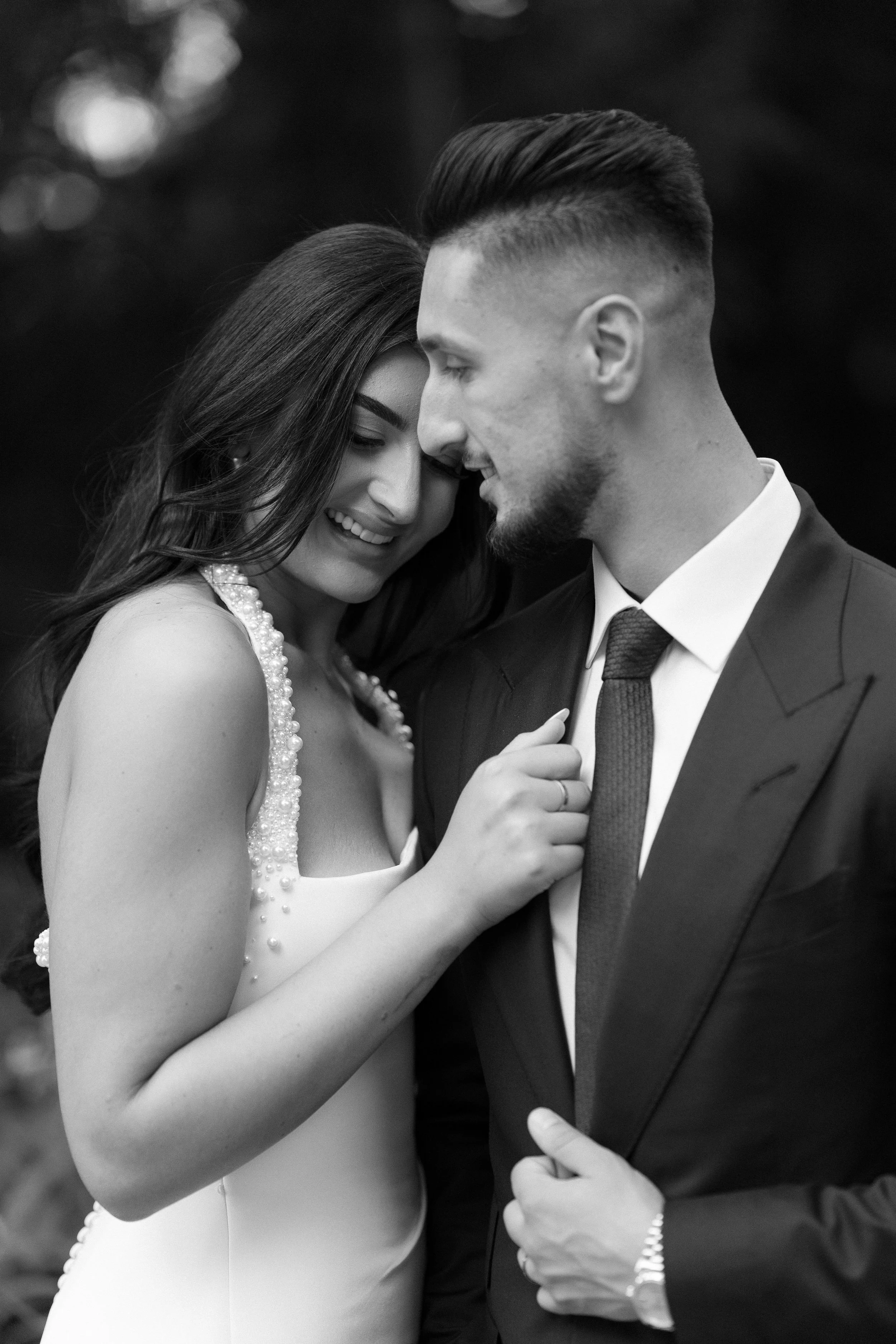 Black and white photo of a smiling couple in wedding attire; the woman has long hair and is wearing a dress with pearl embellishments, and the man has a beard and short hair, wearing a suit and tie, standing closely with their faces almost touching, 