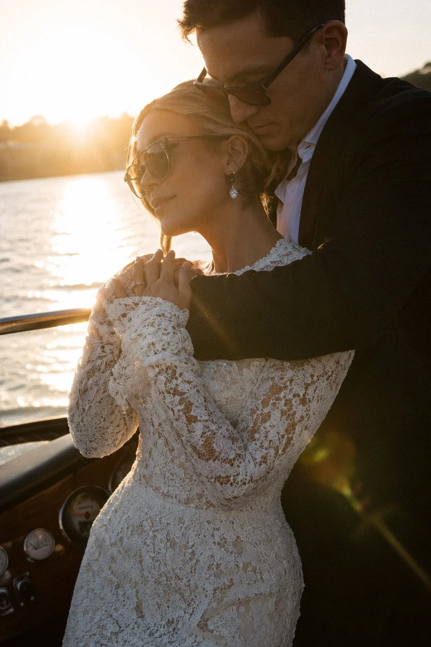 A couple dressed in formal attire embracing on a boat during sunset, with water and shoreline in the background.