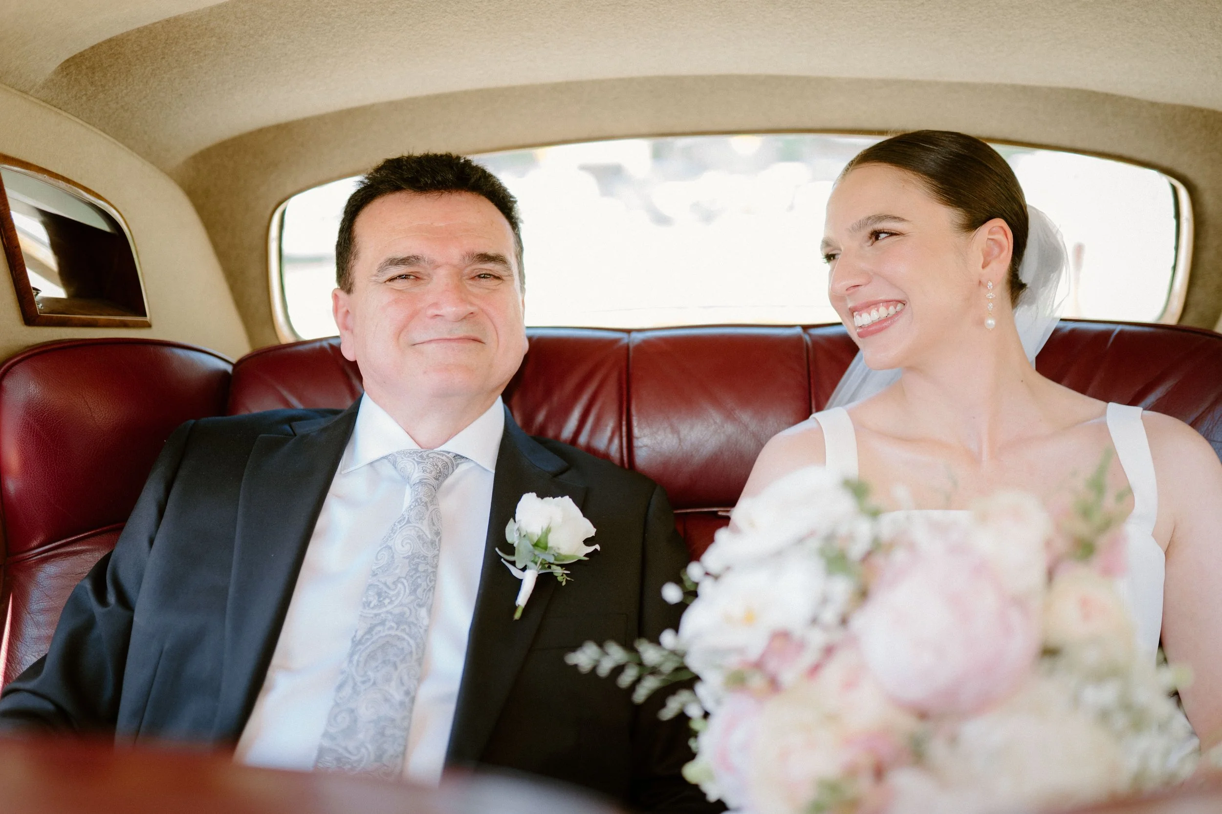 A newlywed couple sitting in the back of a vintage car with red leather seats, the groom in a black tuxedo and the bride in a white wedding dress holding a bouquet.