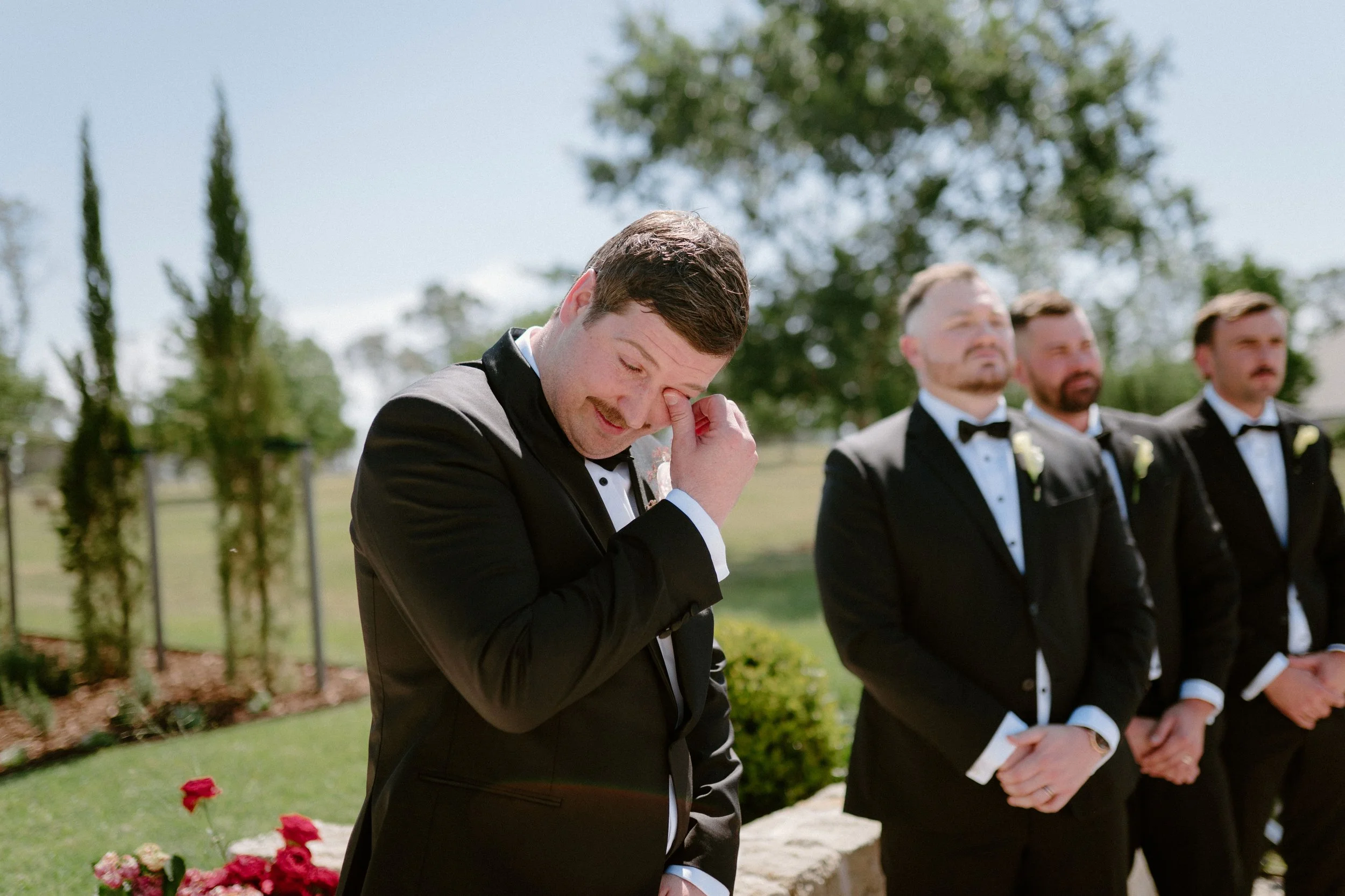 Man in a tuxedo wiping his eye during an outdoor wedding ceremony, with three other men dressed in tuxedos standing behind him.