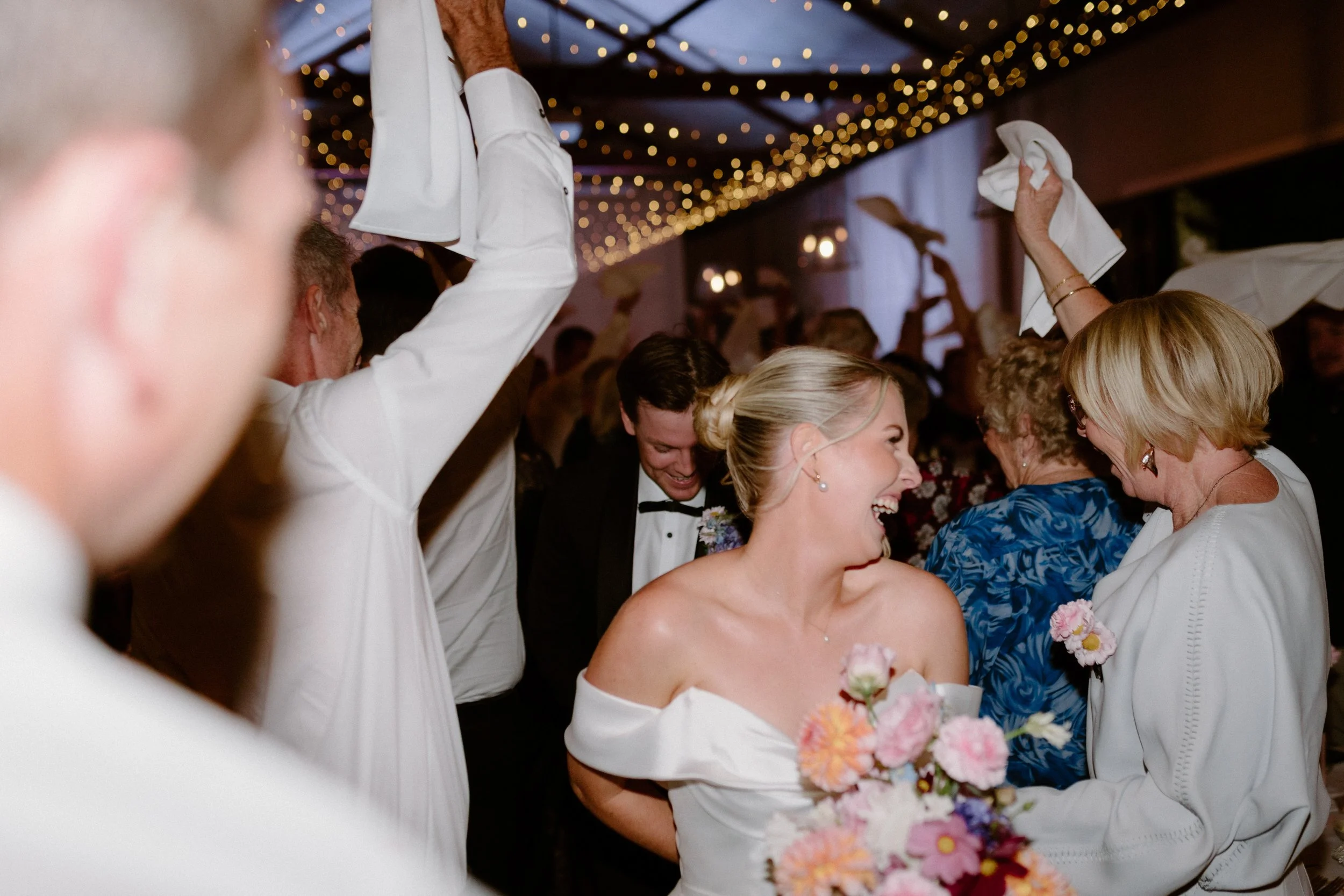 Group of wedding guests at a reception, dancing and celebrating under a ceiling decorated with string lights.