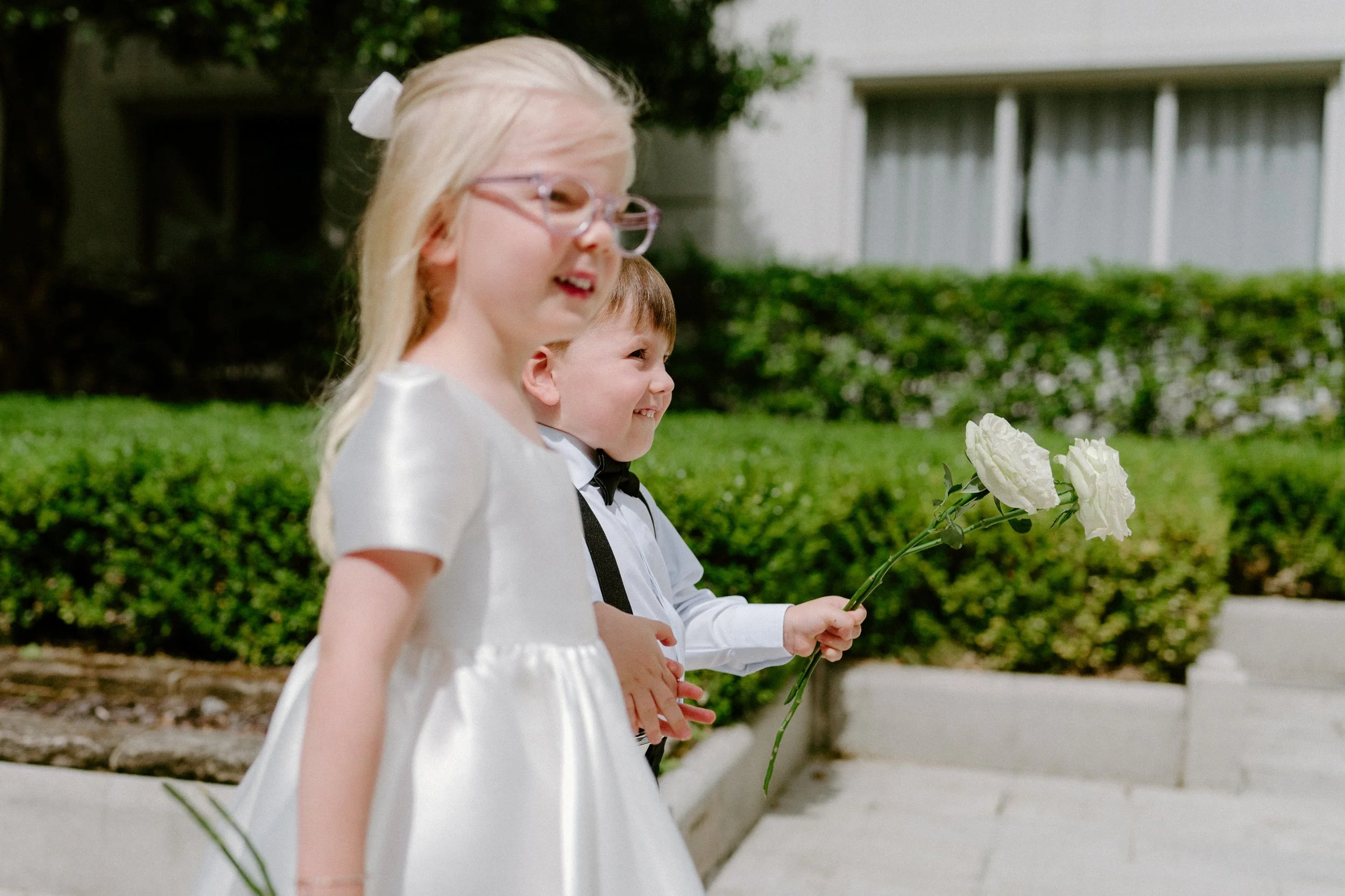Two young children, a girl and a boy, dressed in formal attire, standing outdoors with white flowers in the boy's hand, smiling happily.
