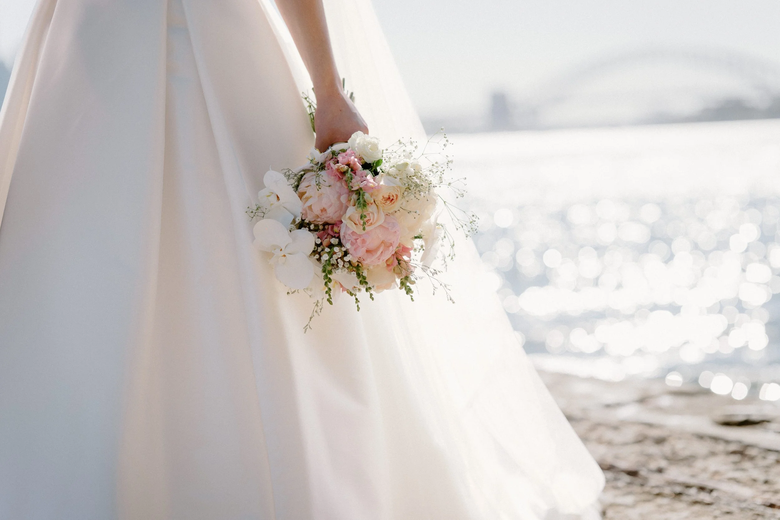 A bride in a white wedding dress holding a bouquet of pink and white flowers on a beach with water and a bridge in the background.