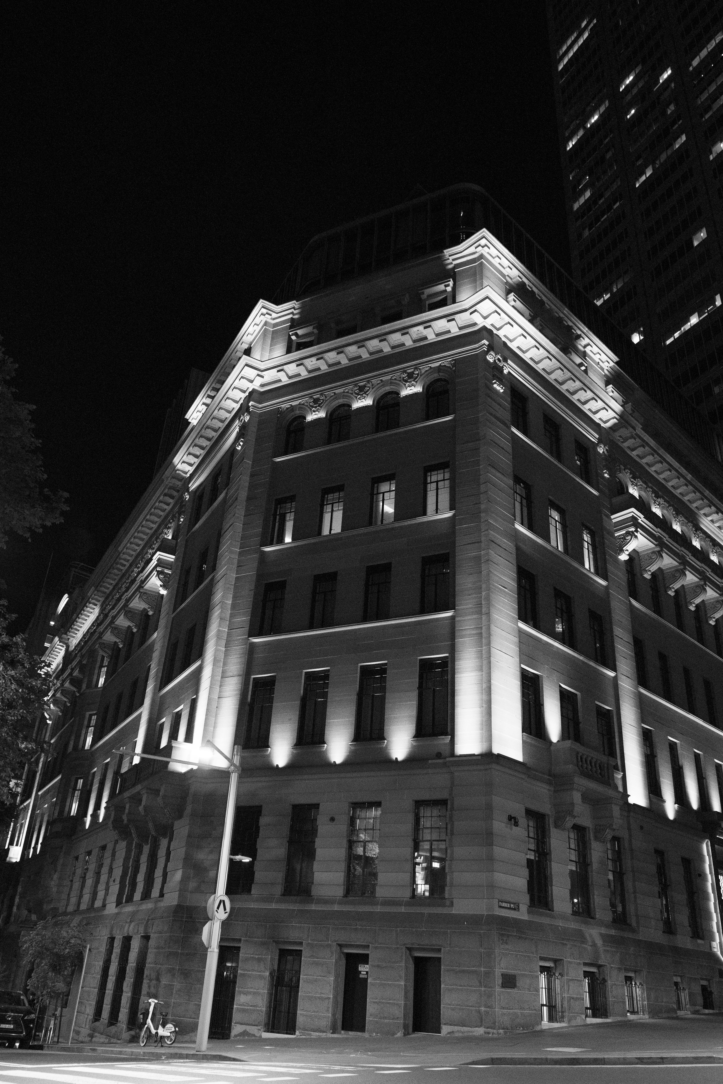 A black and white photo of a well-lit historic building at night on a city street corner, with a modern skyscraper in the background.