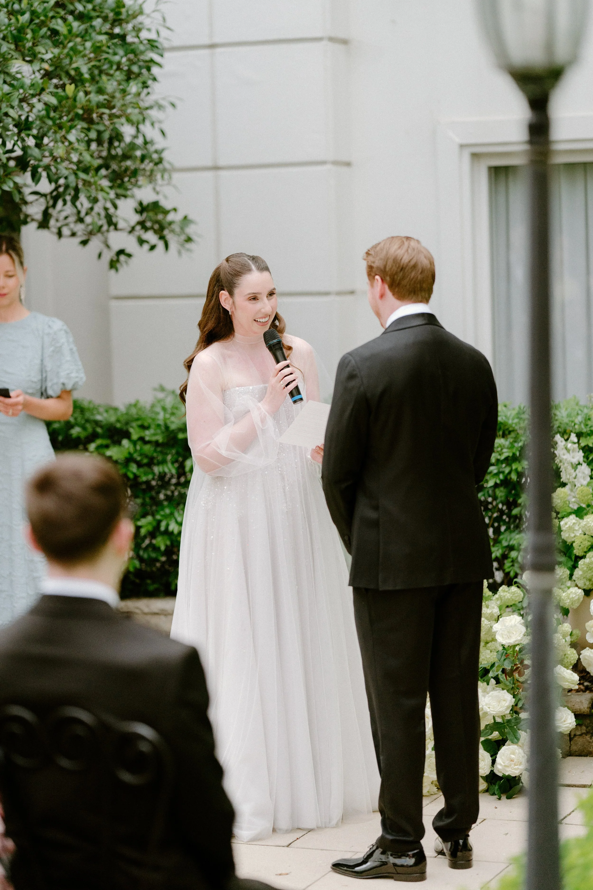 A wedding ceremony outdoors with a bride in a white gown holding a microphone and reading vows, standing in front of a groom in a black suit, with guests seated and a bridesmaid in a lace dress in the background.
