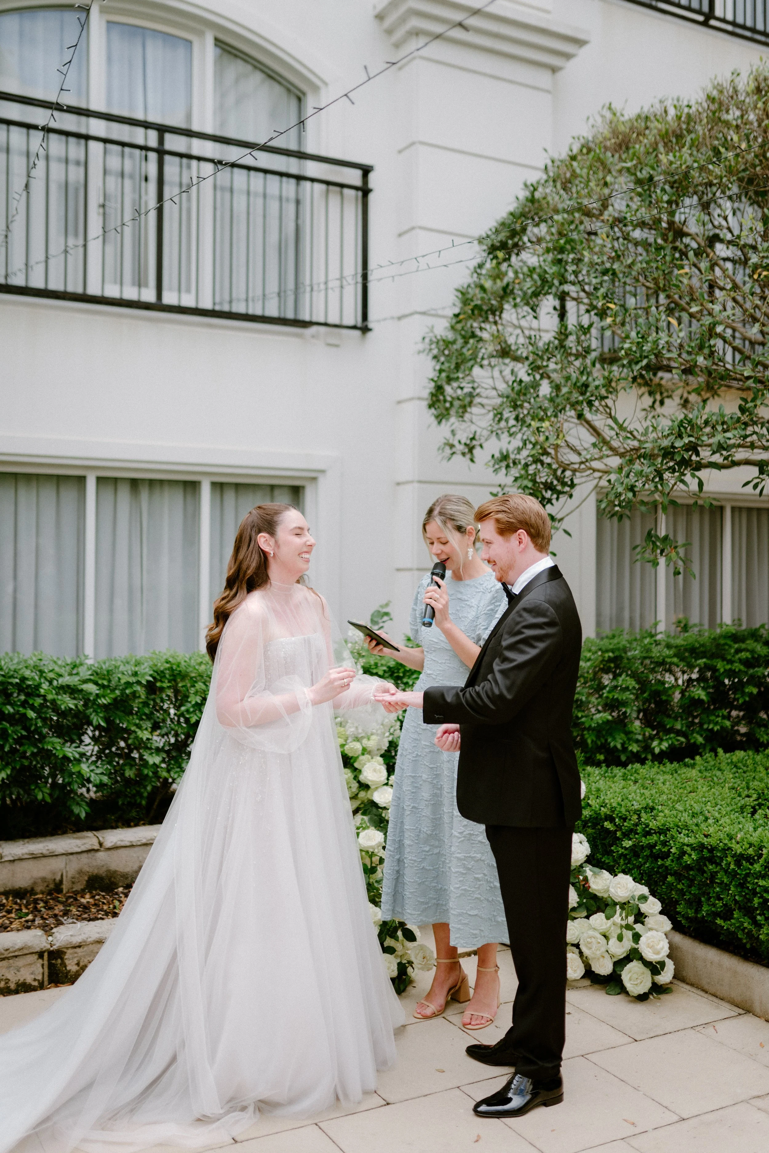 A bride and groom exchanging rings during their outdoor wedding ceremony, with a woman officiant reading from a tablet, all set against a background of greenery and a white building.