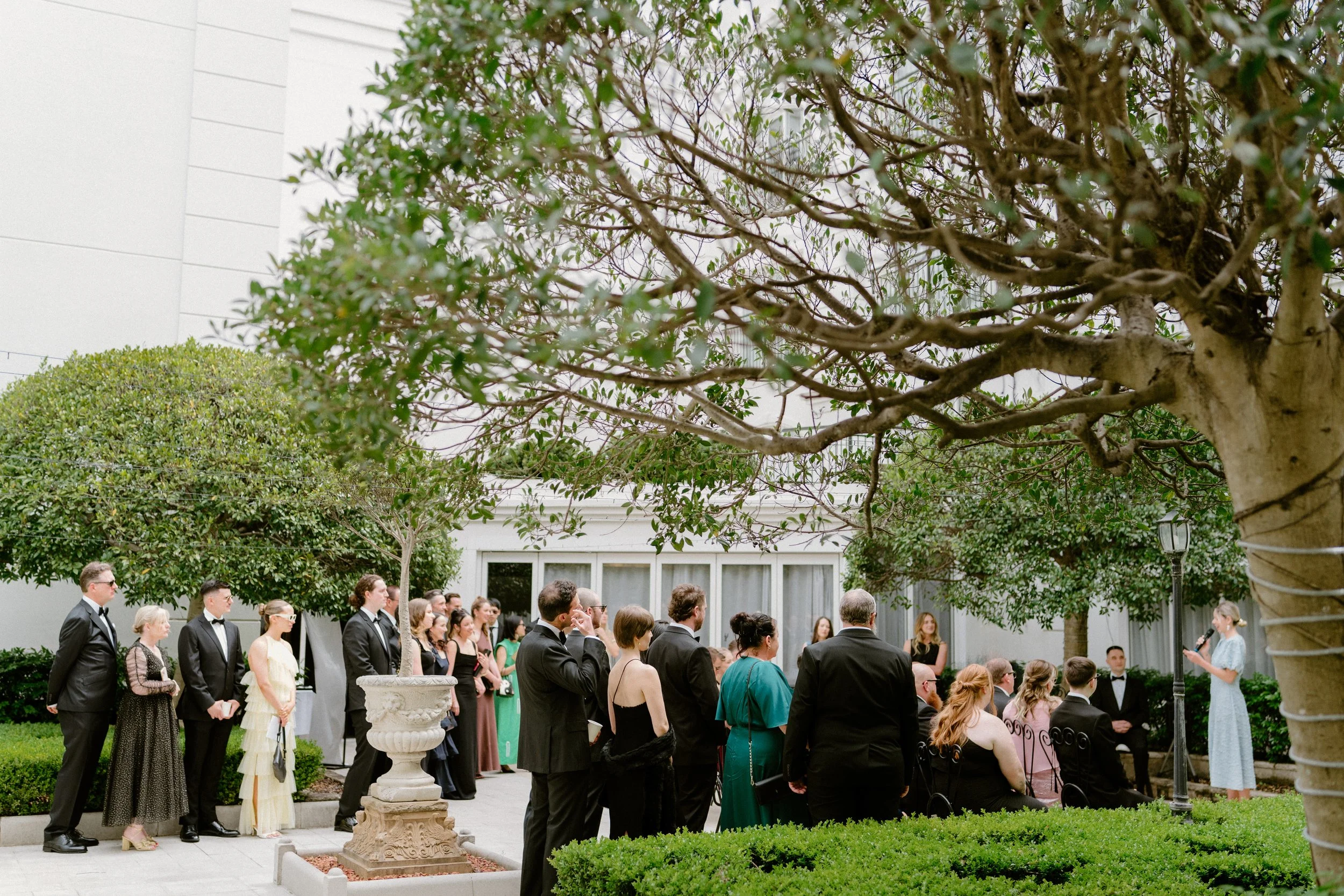 People in formal attire attending an outdoor wedding ceremony, with a woman giving a speech, surrounded by trees and greenery.