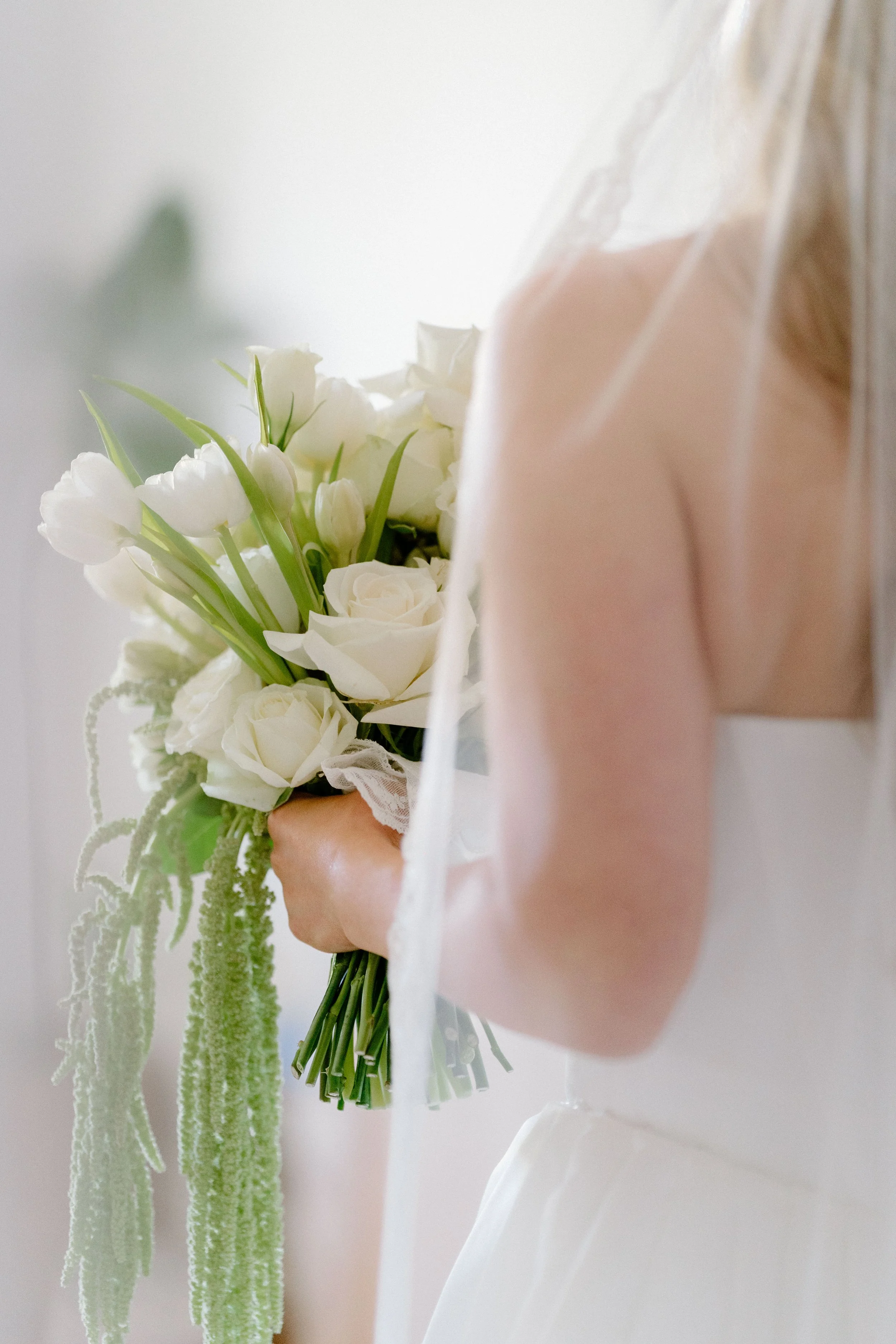 A person holding a bouquet of white roses and tulips with green leaves and stems, standing near a wall with a tall green plant in the background.