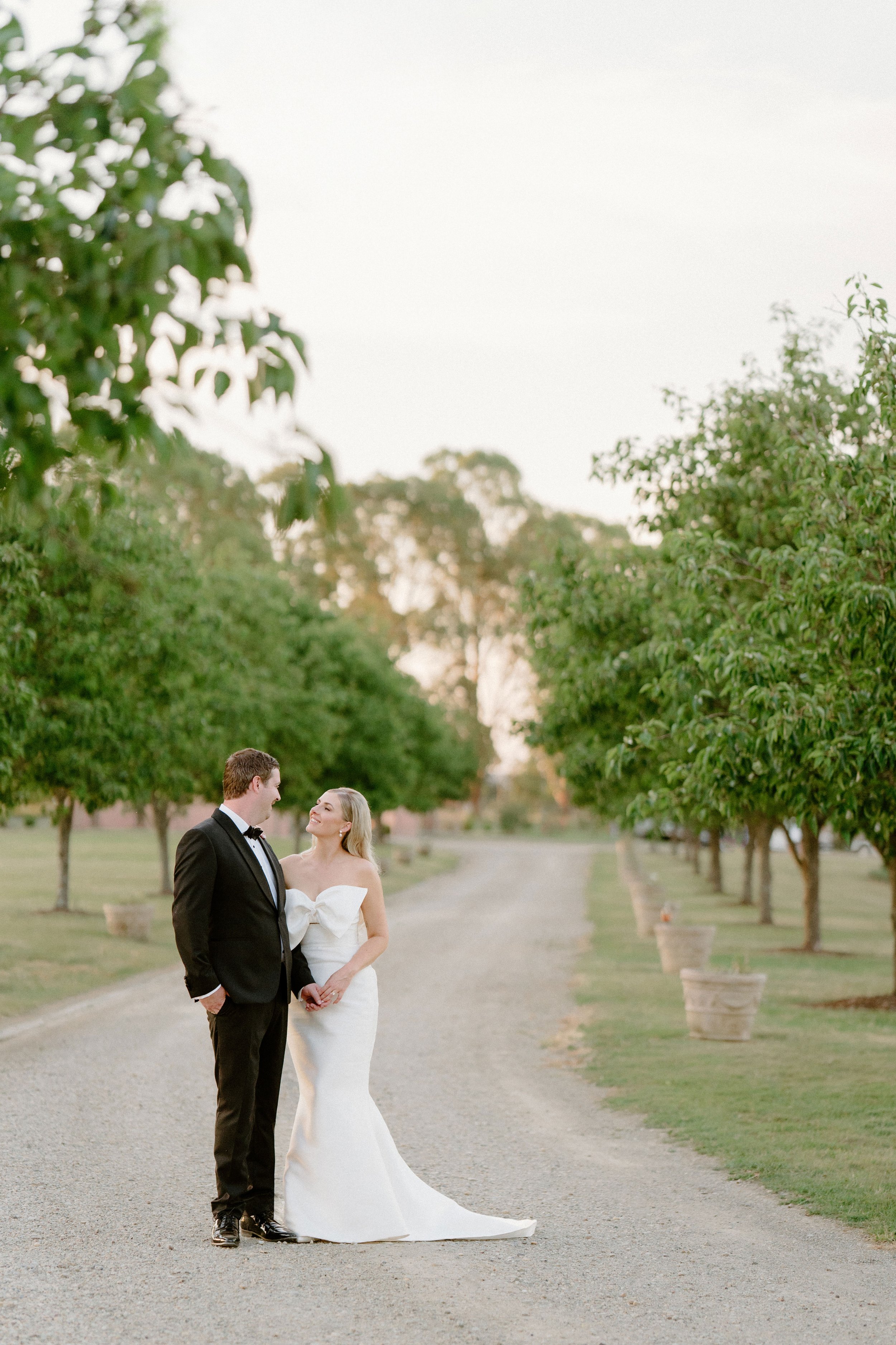 The Sienna, Hunter Valley Wedding Venue, Wedding Photos. A bride and groom holding hands and smiling at each other on a gravel pathway lined with green trees during sunset.