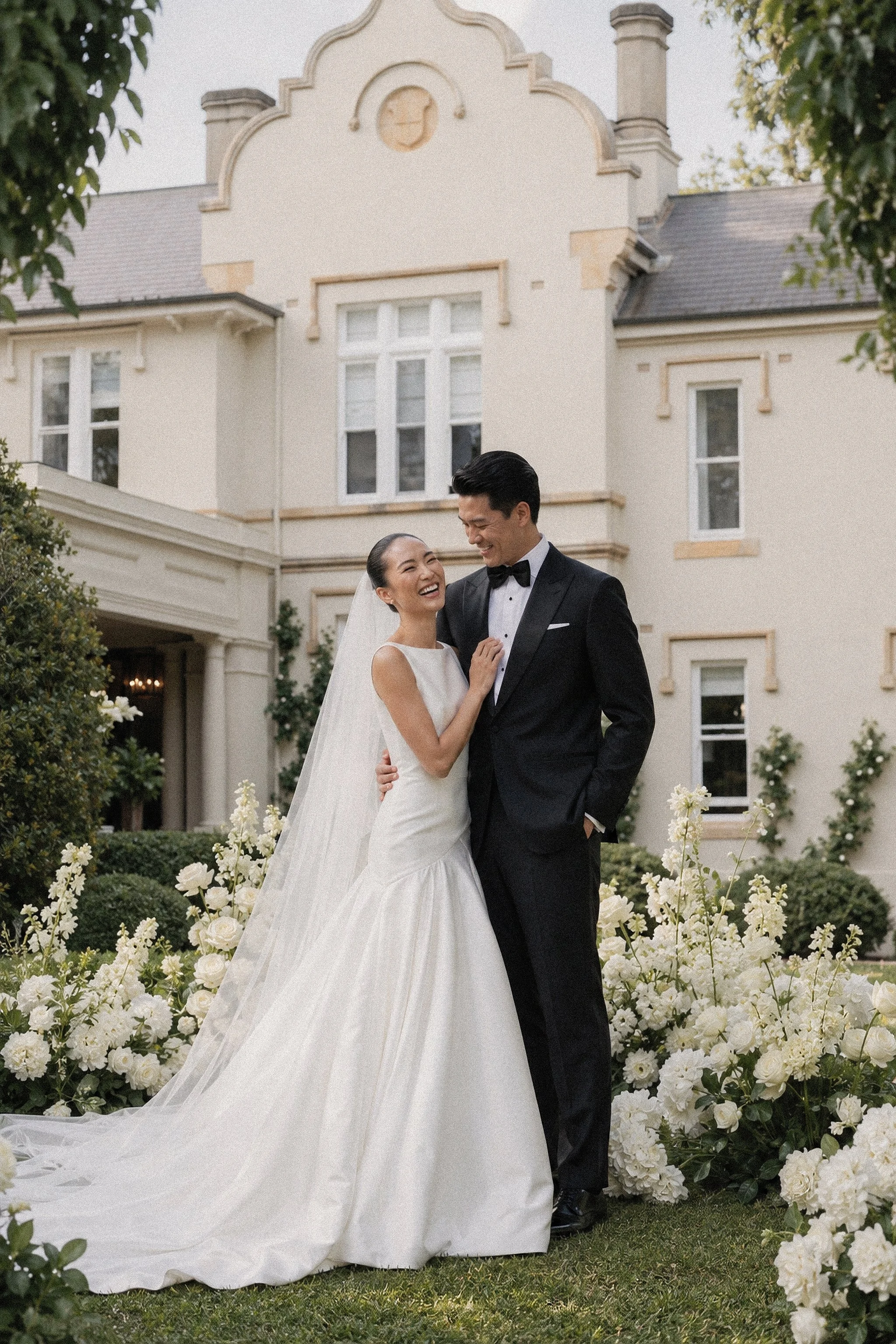 A newlywed couple, a woman in a white wedding gown and a man in a black tuxedo, share a joyful moment in front of a large, elegant house surrounded by white flowers and greenery.
