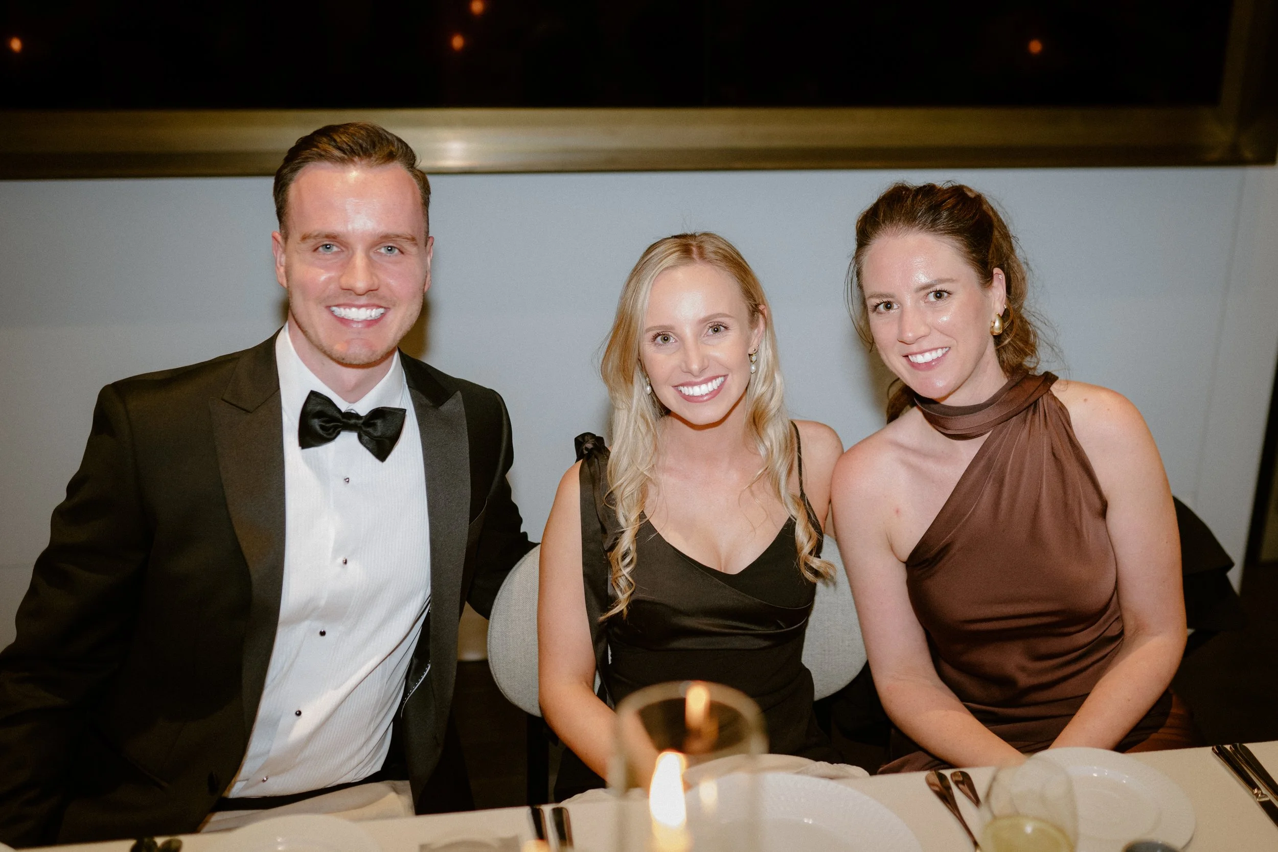 Three people sitting at a formal dinner table, dressed in elegant attire, smiling at the camera during an evening event.