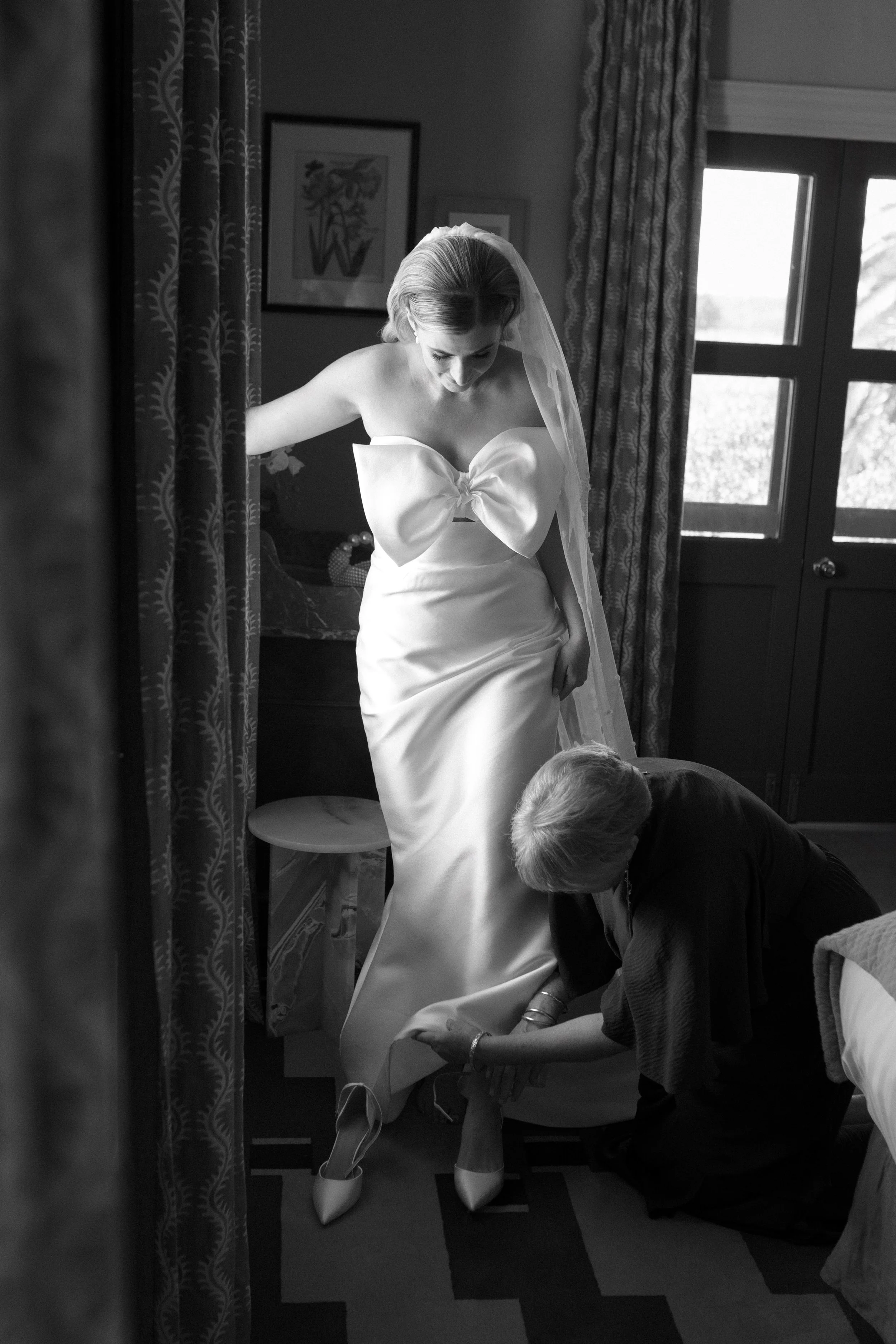 A bride in a wedding dress with a large bow on the front is being assisted with her shoes by another woman in a dimly lit room, with curtains and framed pictures on the wall behind them.