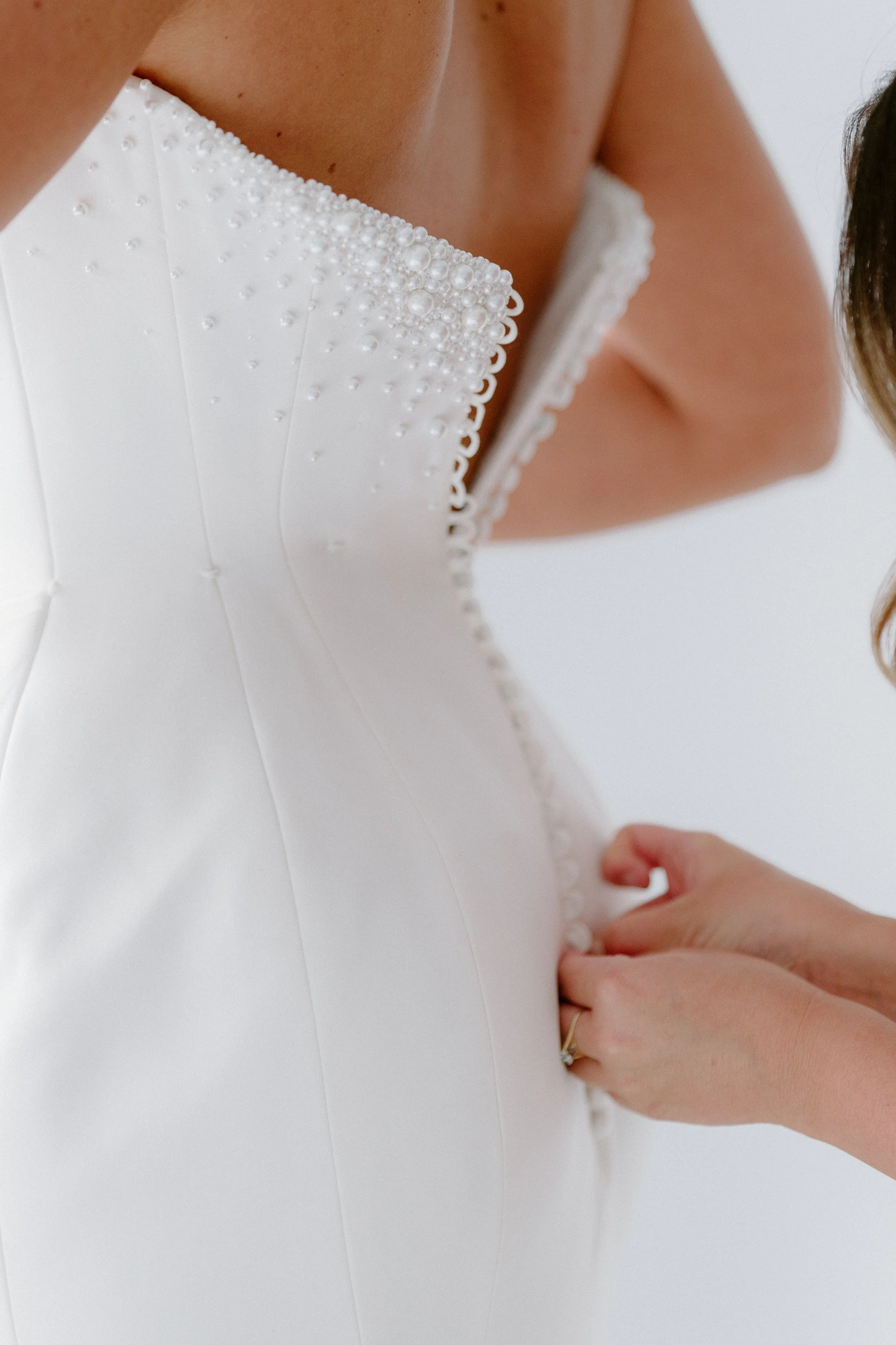 Close-up of a bride's white wedding dress with pearl embellishments, being fastened at the back by another person.