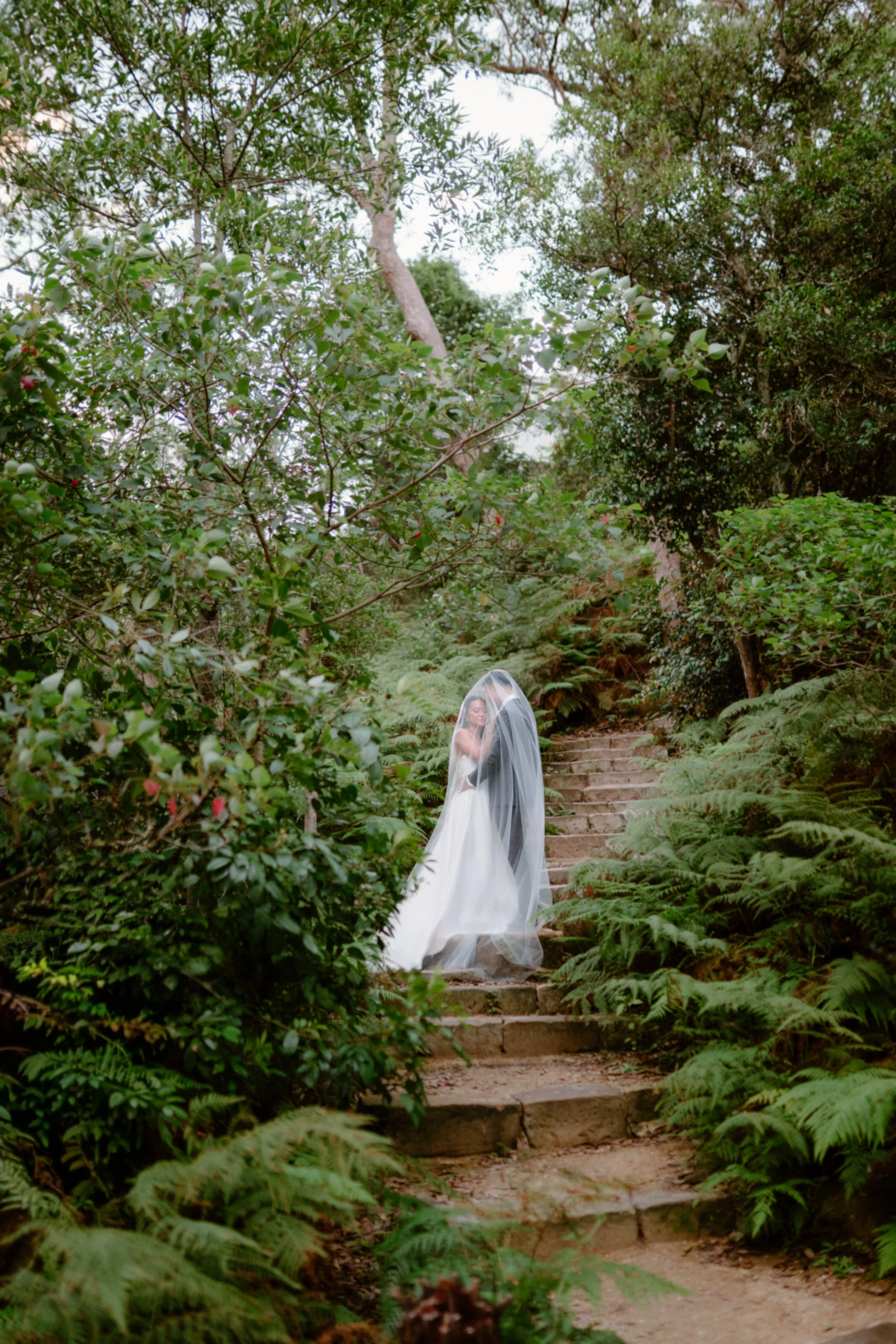A bride in a wedding dress and veil walking up stone steps in a lush green forest.