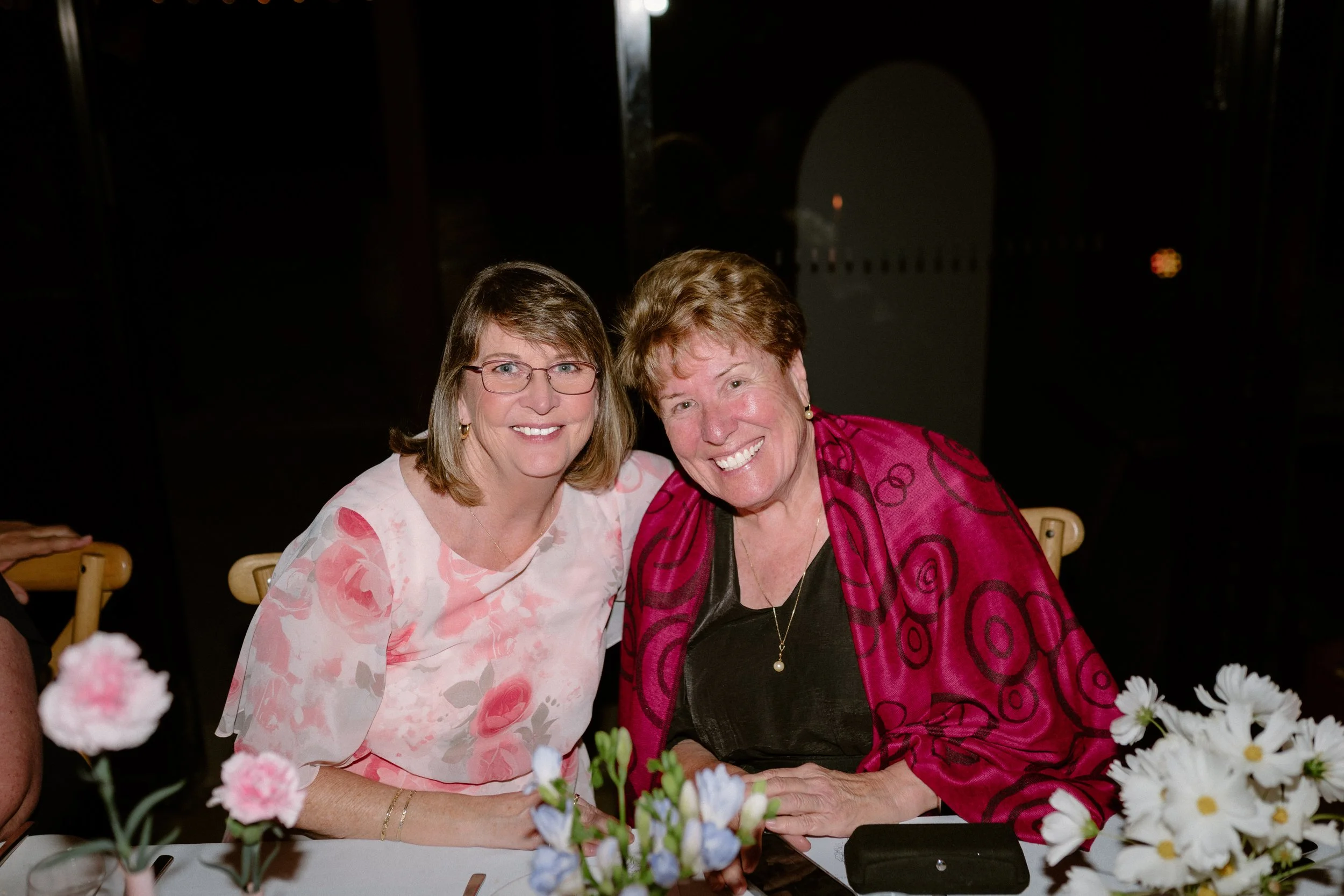 Two women sitting at a table, smiling, with flowers in the foreground. One woman wears glasses and a light-colored dress with pink and red patterns, the other wears a pink and black patterned jacket.