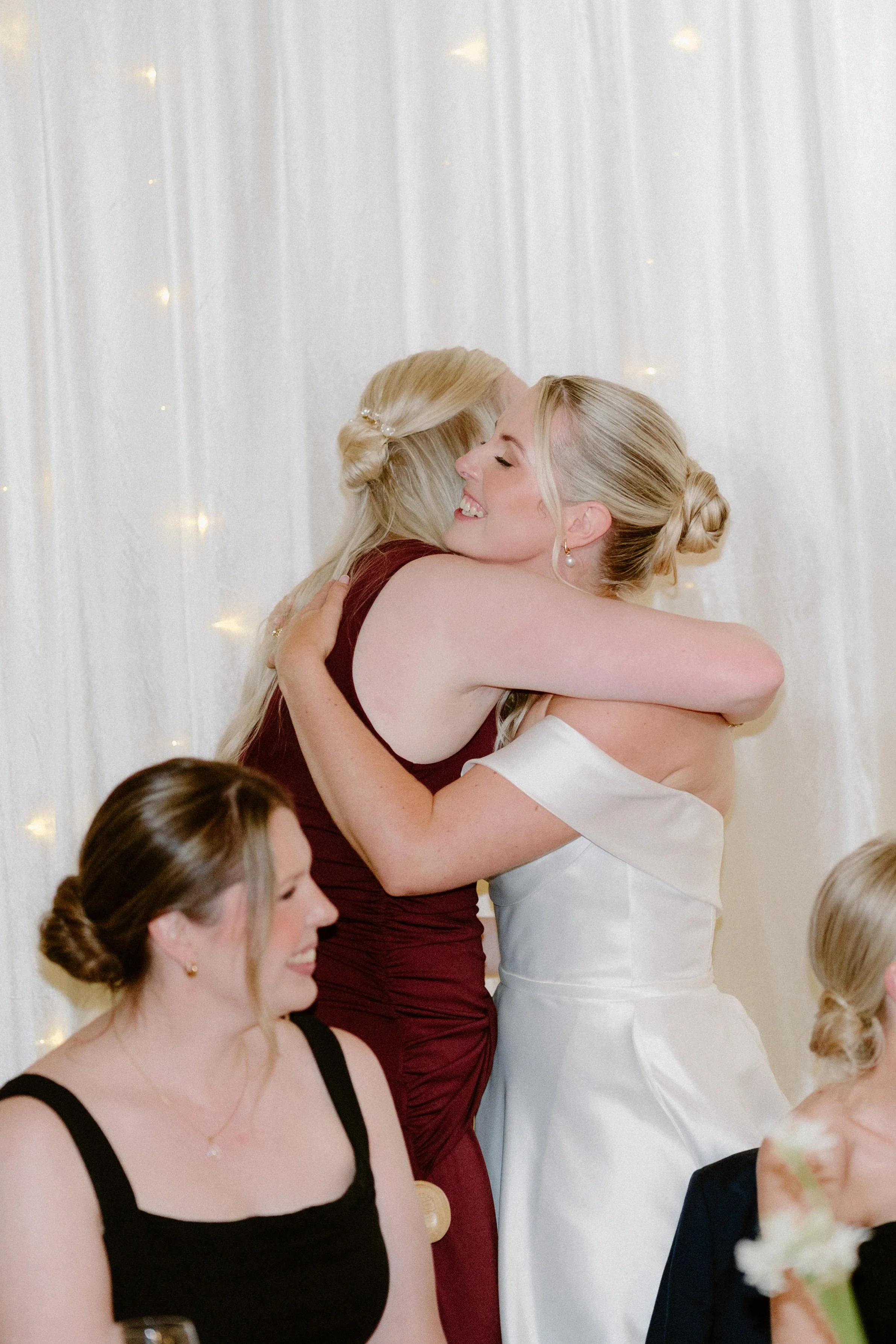 Two women hugging and smiling at a wedding reception. The woman in the white dress is the bride, and the woman in the maroon dress is a guest.