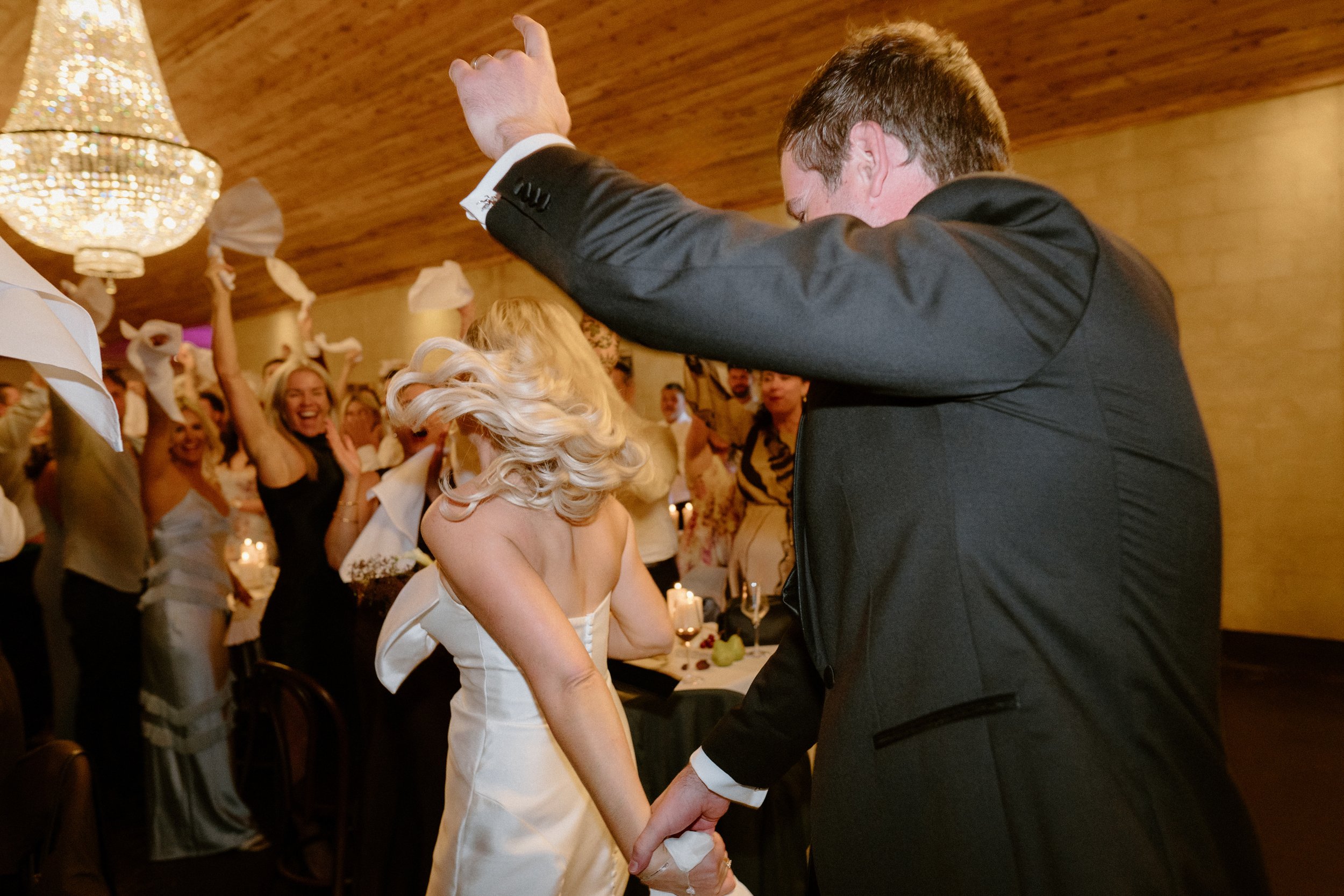 A wedding reception with a bride and groom dancing, surrounded by joyful guests holding napkins in the air.