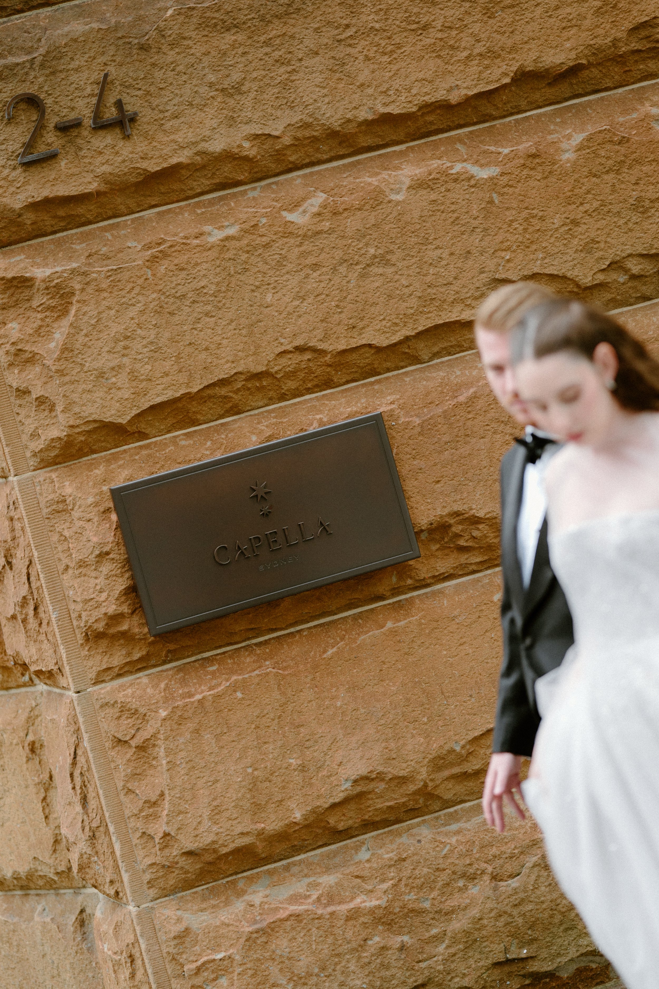 Close-up of a brown stone building with the address 2-4 and a black plaque that reads 'Capella Sydney'. Part of a bride in a wedding dress and a groom in a tuxedo are visible in the foreground.