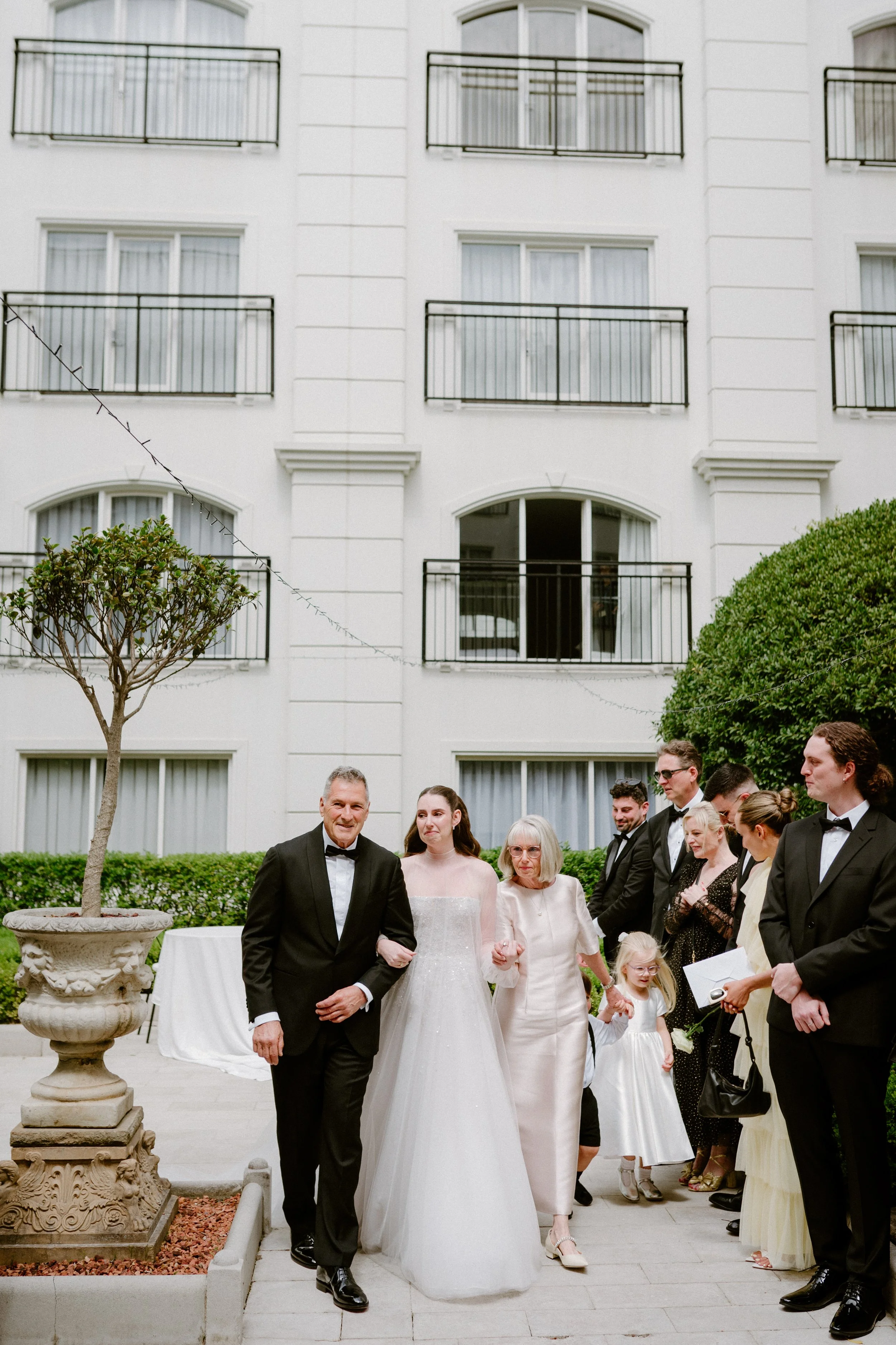 A group of people in formal attire, including a bride in a white wedding gown, is gathered outside a white building with balconies, for a wedding celebration.