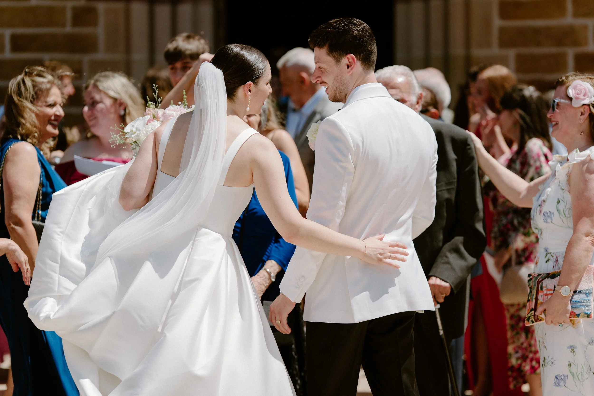 Bride and groom smiling and holding hands at their wedding reception, surrounded by guests.