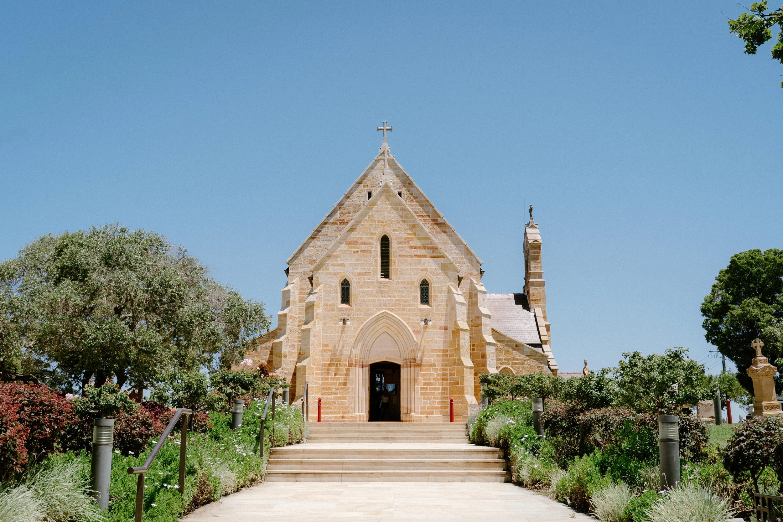 A stone church with a cross on top, surrounded by greenery and flowers on a clear, sunny day.