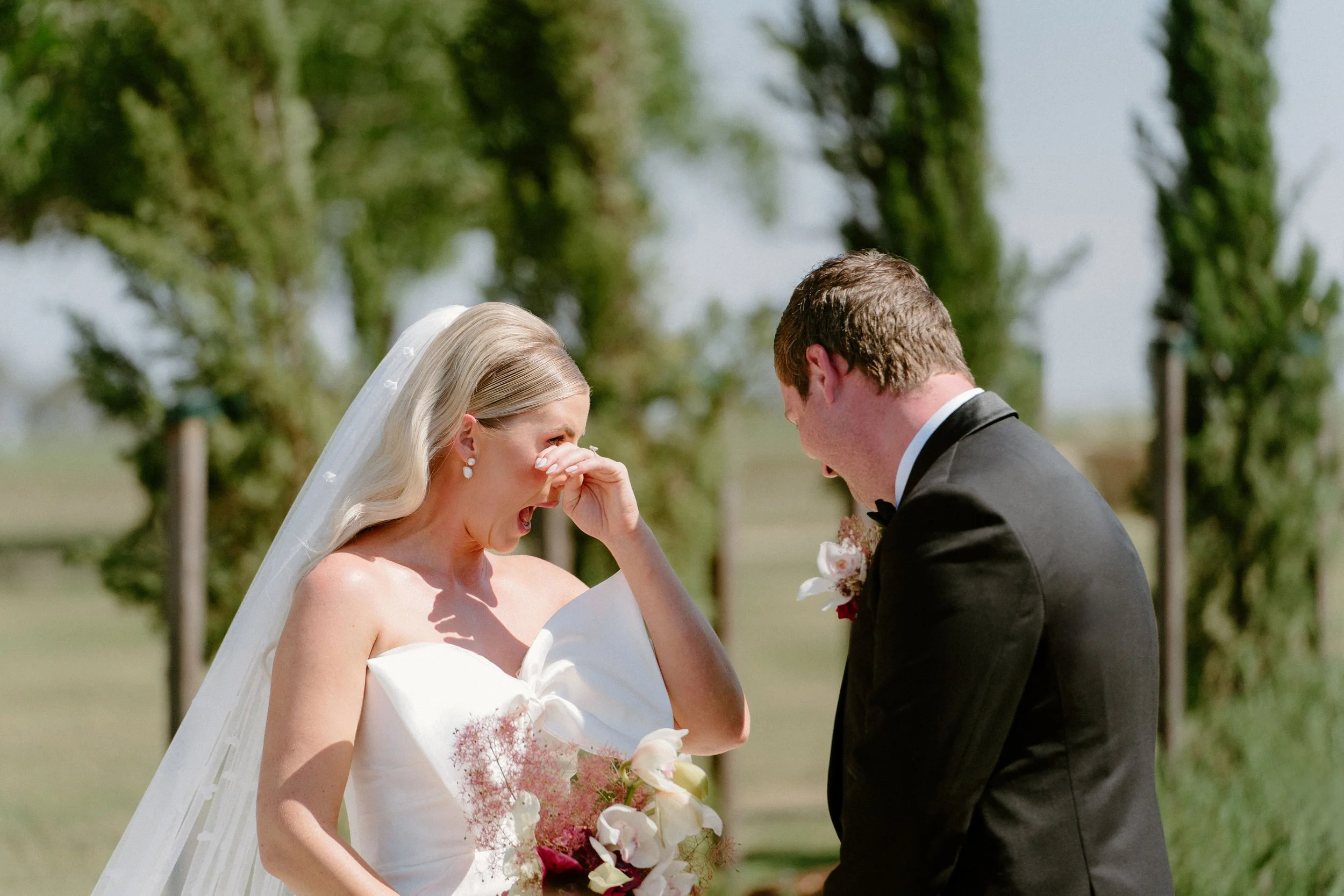 A bride and groom are sharing a joyful moment outside on their wedding day. The bride is wearing a strapless white wedding gown and veil, holding a bouquet of flowers. She is wiping away tears of happiness as she looks at her groom. The groom is dres