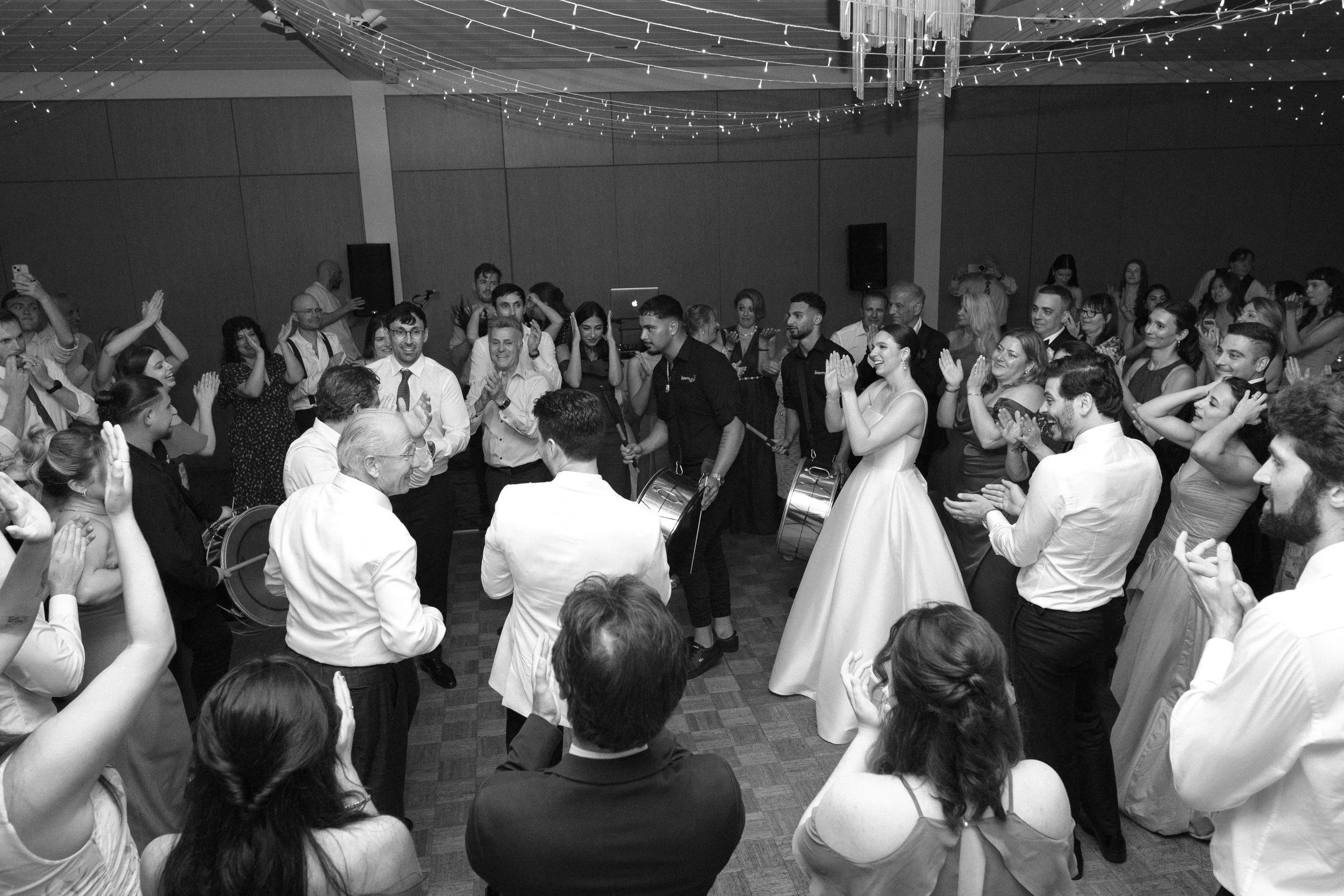 People dancing and celebrating at a wedding reception in a decorated hall, with some playing drums and others clapping and enjoying the moment.