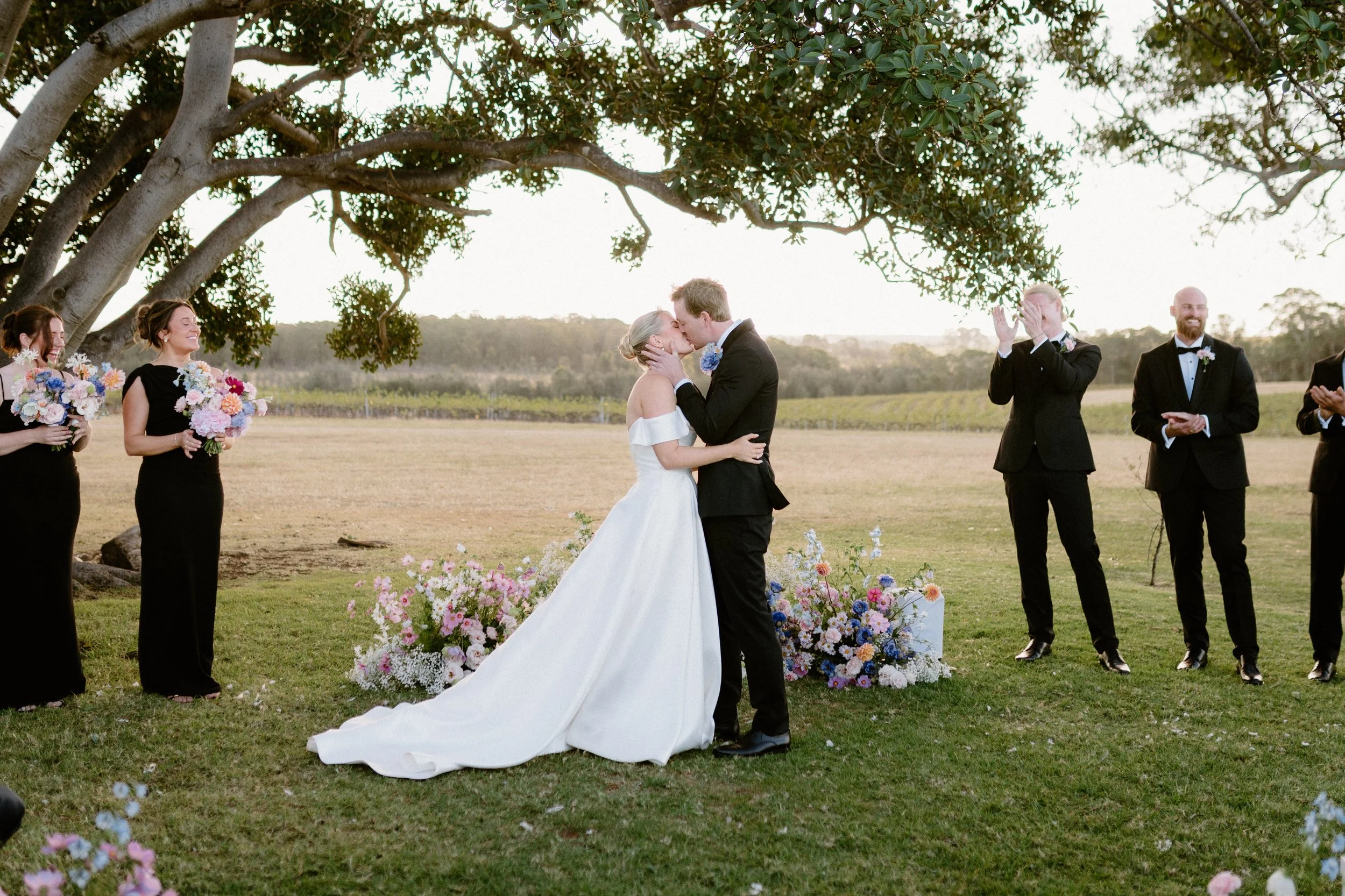 A bride and groom share a kiss during an outdoor wedding ceremony under a tree with officiants and bridesmaids holding bouquets watching nearby. Bimbadgen Palmers Lane Wedding, Hunter Valley