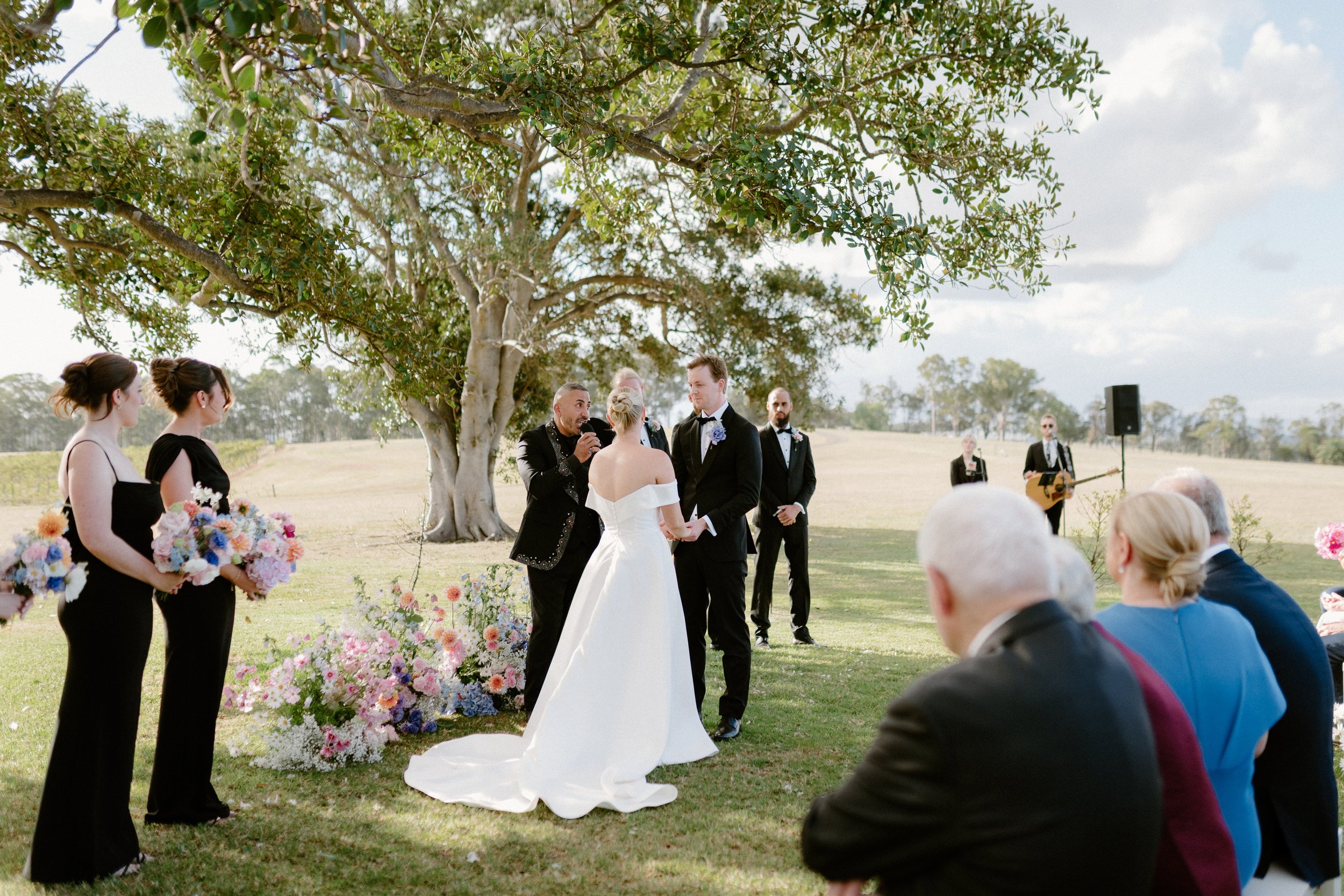 A wedding ceremony outdoors with a bride and groom exchanging vows under a large leafy tree. Bridesmaids hold colorful bouquets, and groomsmen stand nearby. Guests are seated and watching the ceremony, with some music performers in the background. 